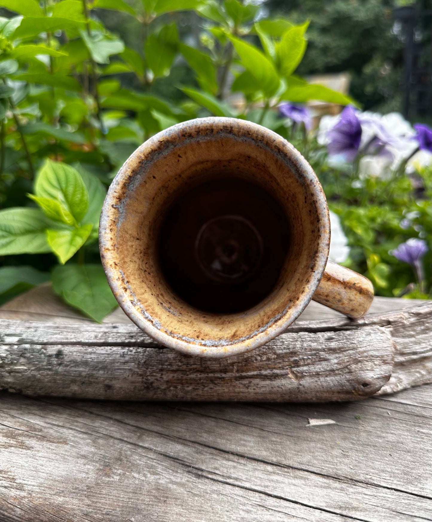 A handmade ceramic mug with a rustic, speckled brown and beige glaze, shown from a top-down perspective resting on a weathered piece of driftwood in an outdoor garden setting.