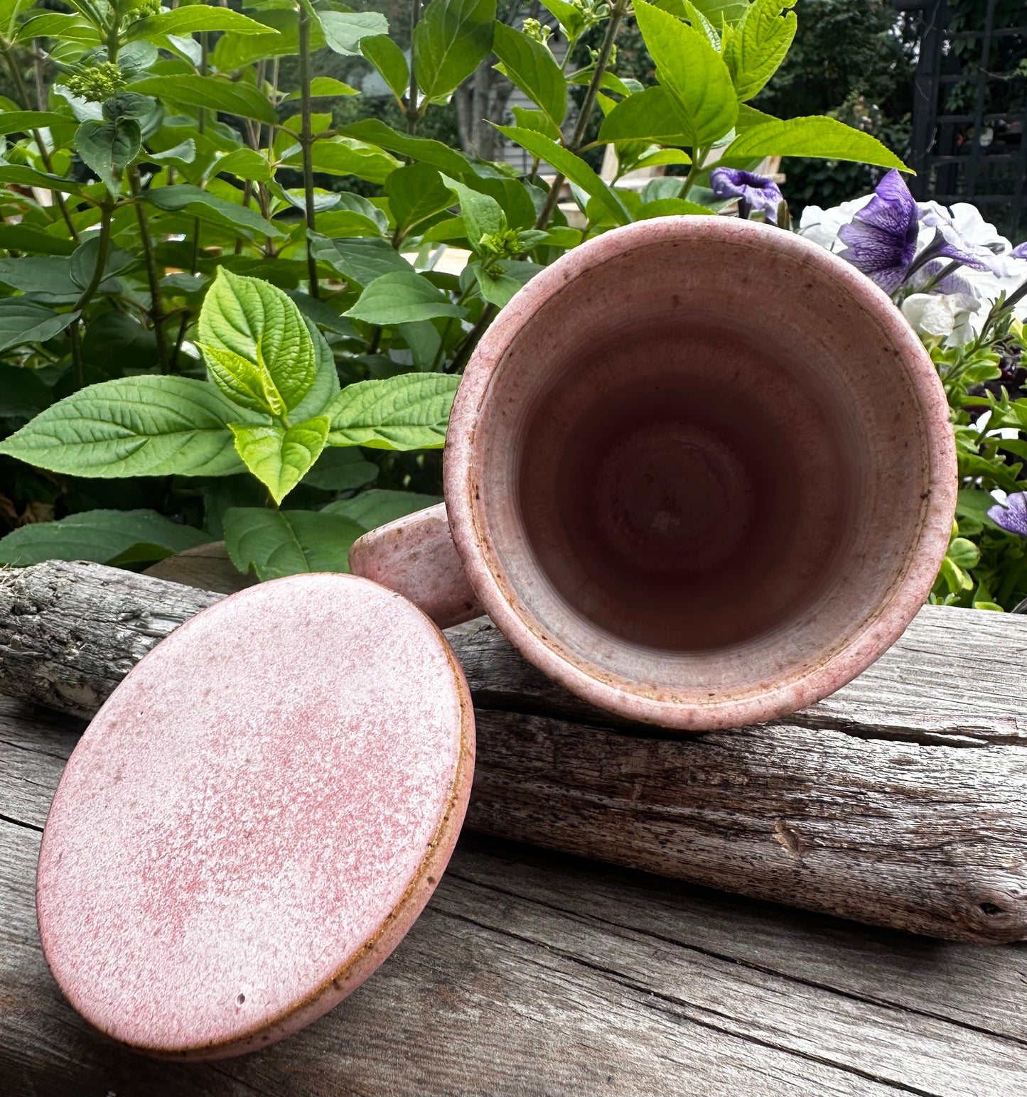 A handmade pink glazed ceramic travel mug with its matching lid resting on a weathered log outdoors, with green garden foliage and purple flowers in the background.