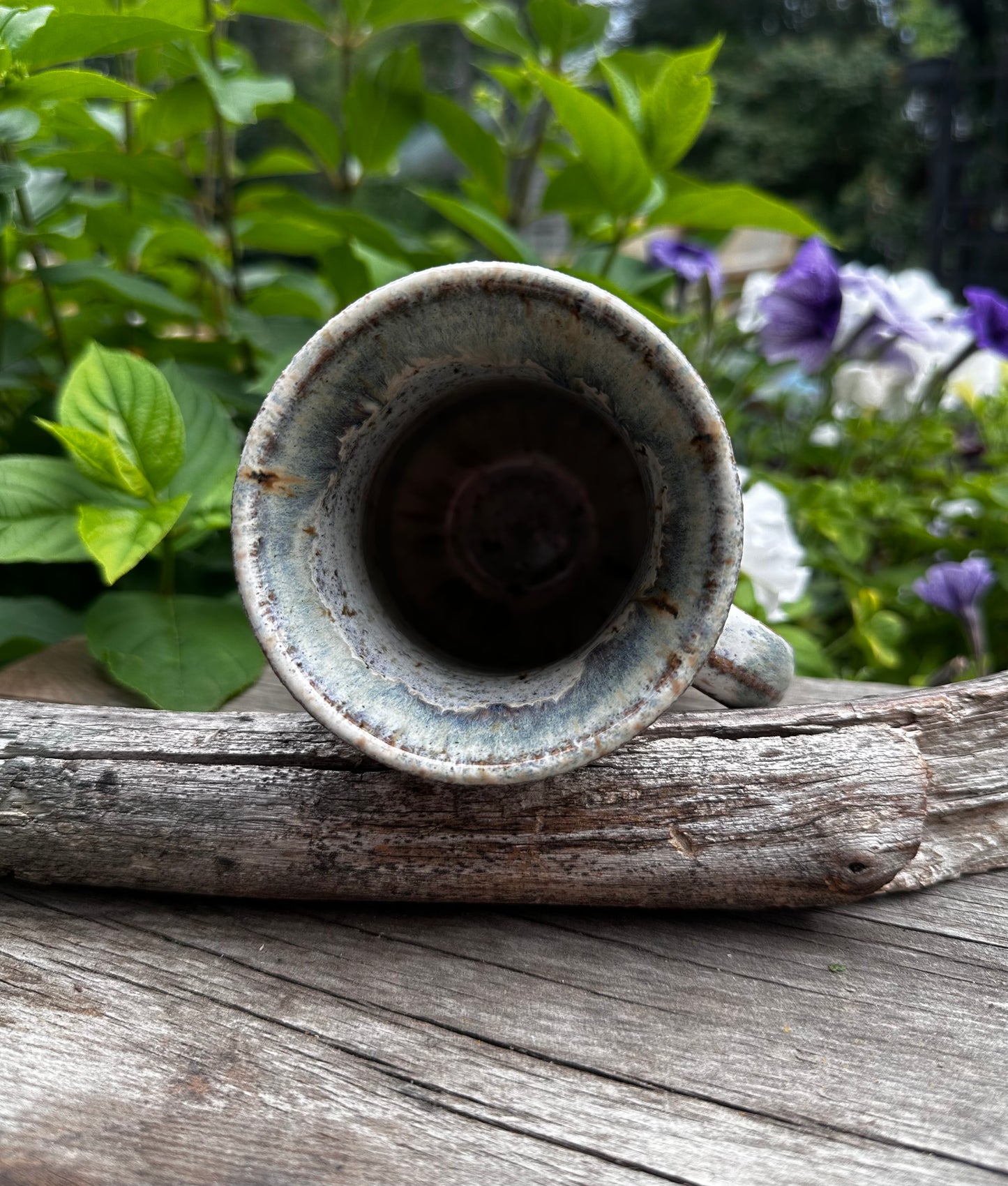 A rustic ceramic mug with a distressed blue-white glaze, resting on a weathered log with green garden foliage and purple flowers in the background.