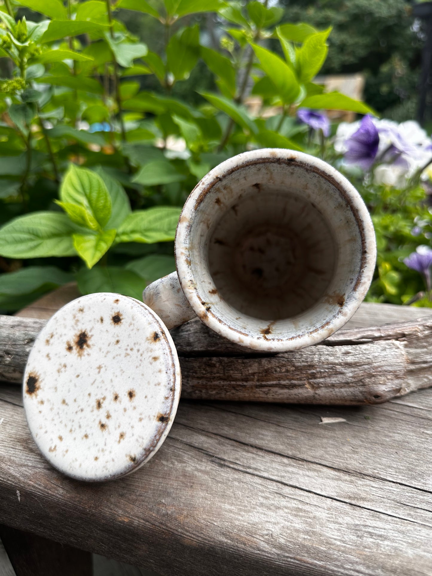 A handmade ceramic travel mug with lid, both featuring a speckled off-white glaze with dark brown spots, shown outdoors against a weathered wooden post and green foliage.