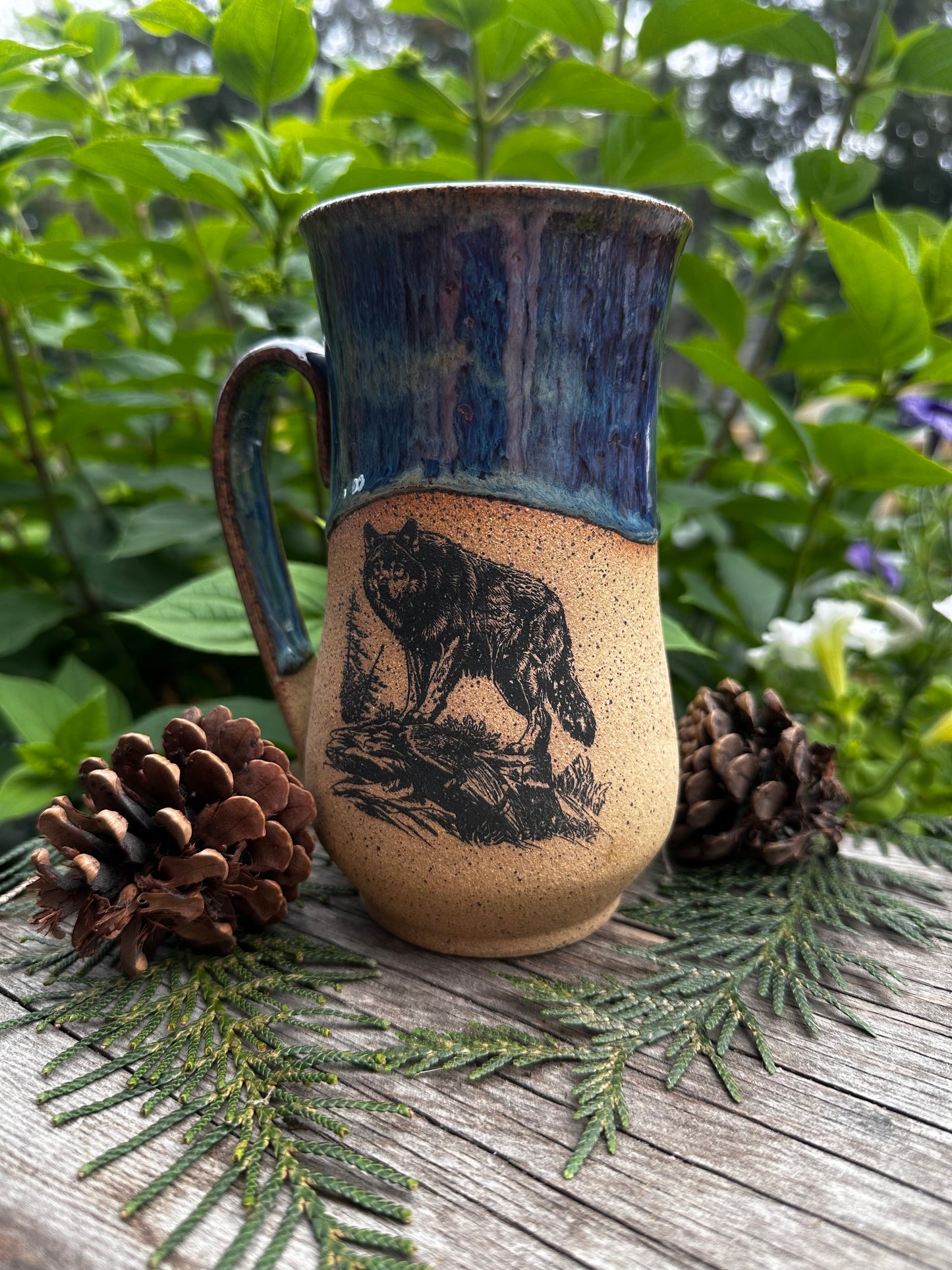 A handmade ceramic mug with a rustic design, featuring a dark blue glazed handle and rim, and a speckled beige body with a black wolf illustration. The mug is styled on a wooden surface with pine needles and pinecones, set against a background of green foliage.