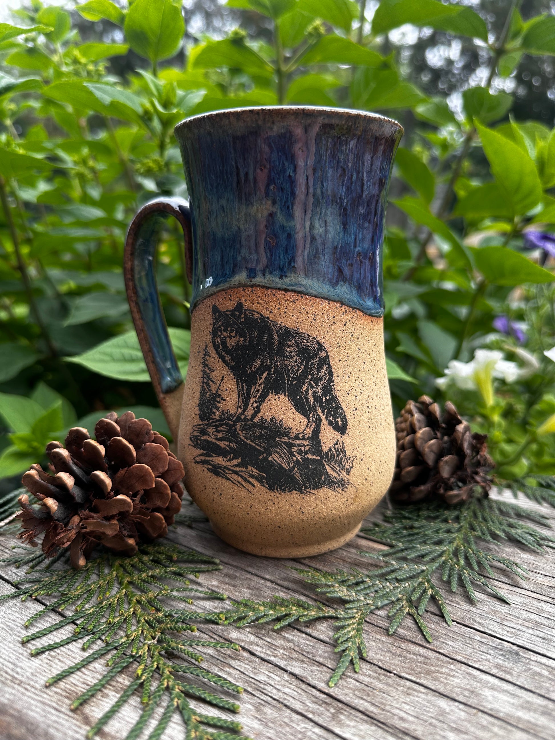 A handmade ceramic mug with a rustic design, featuring a dark blue glazed handle and rim, and a speckled beige body with a black wolf illustration. The mug is styled on a wooden surface with pine needles and pinecones, set against a background of green foliage.