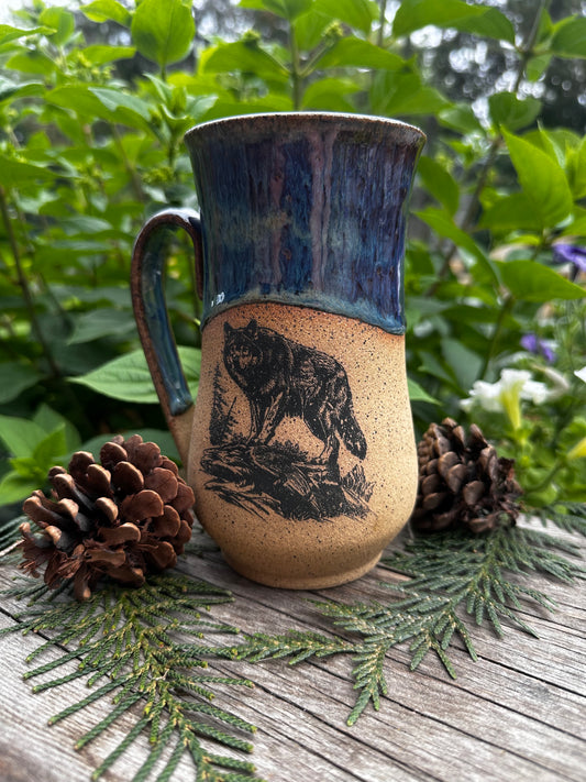 A handmade ceramic mug with a rustic design, featuring a dark blue glazed handle and rim, and a speckled beige body with a black wolf illustration. The mug is styled on a wooden surface with pine needles and pinecones, set against a background of green foliage.