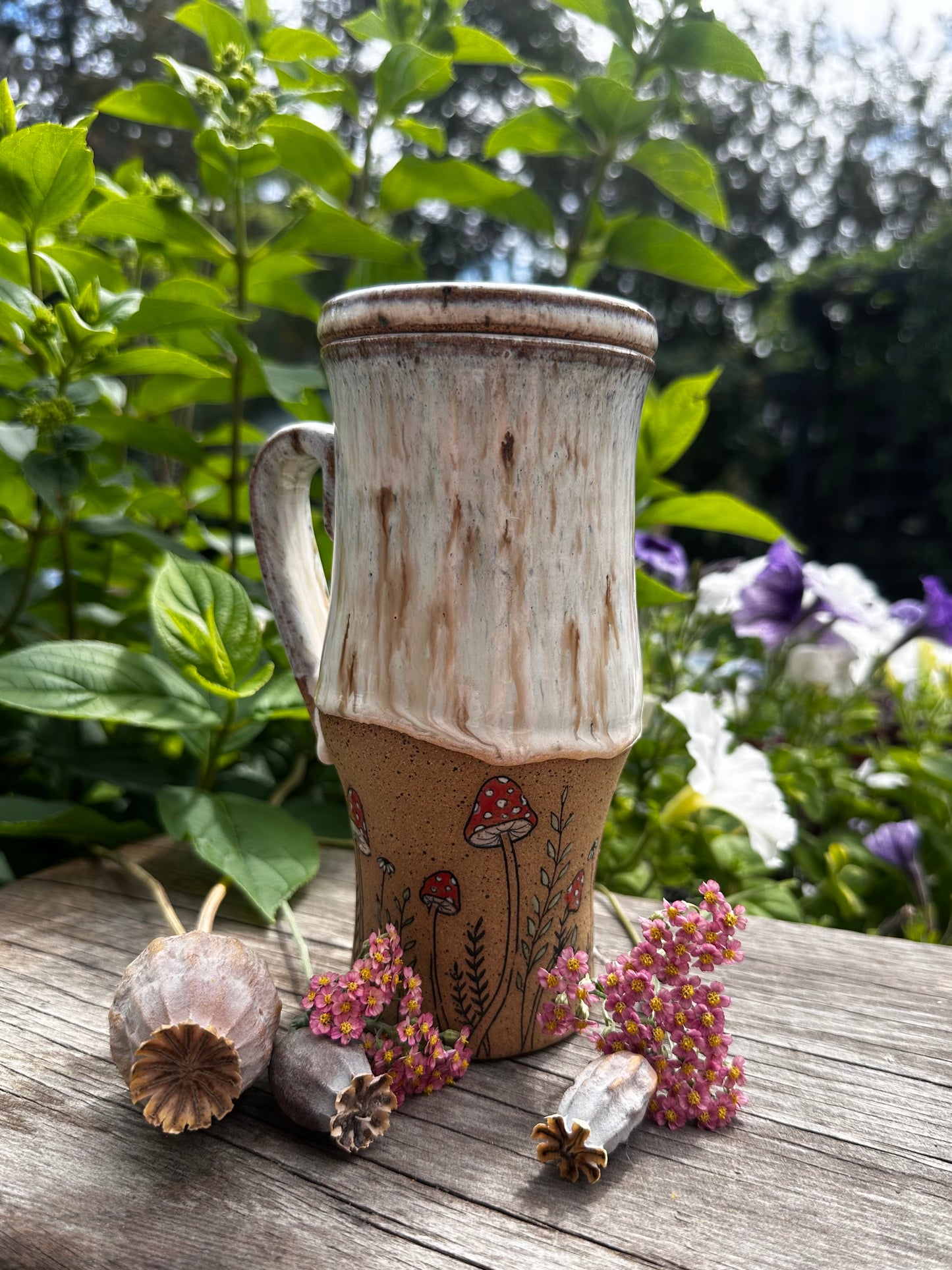 A handmade ceramic travel mug with a rustic, distressed glaze sits on a weathered wooden surface outdoors, surrounded by green foliage and small dried flowers. The mug features a cream-colored top section with brown speckling and a tan-brown bottom section decorated with a hand-painted red and black mushroom motif.