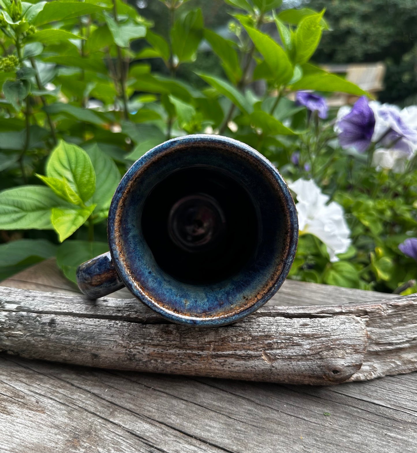 A handmade ceramic mug with a dark blue and brown glaze, shown from a top-down view resting on a weathered piece of wood with a garden background.