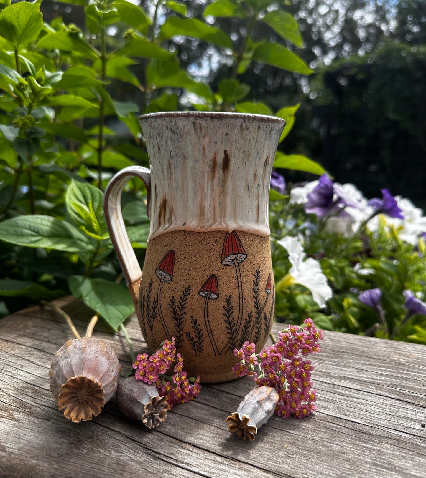 A handmade ceramic mug with a rustic handle, featuring a speckled cream upper half and a brown lower half decorated with red mushrooms and black stems. It is styled on a weathered wooden table with dried flowers and poppy pods, set against a soft-focus background of green garden foliage and purple flowers.