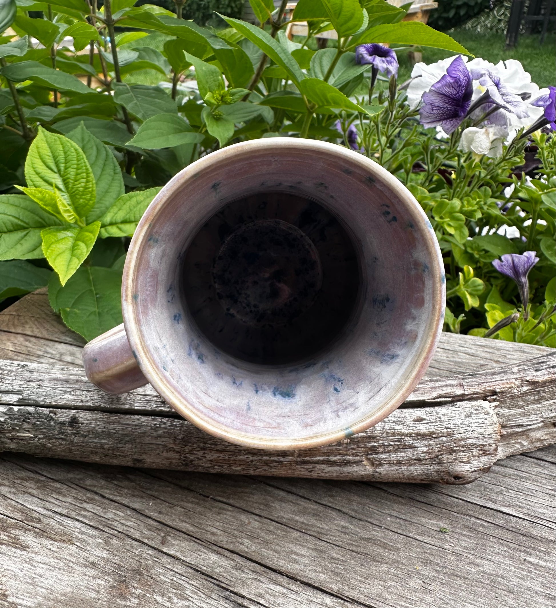 A handmade ceramic mug with a speckled lavender and blue glaze, shown from a top-down perspective resting on a weathered piece of wood with green garden foliage and purple flowers in the background.