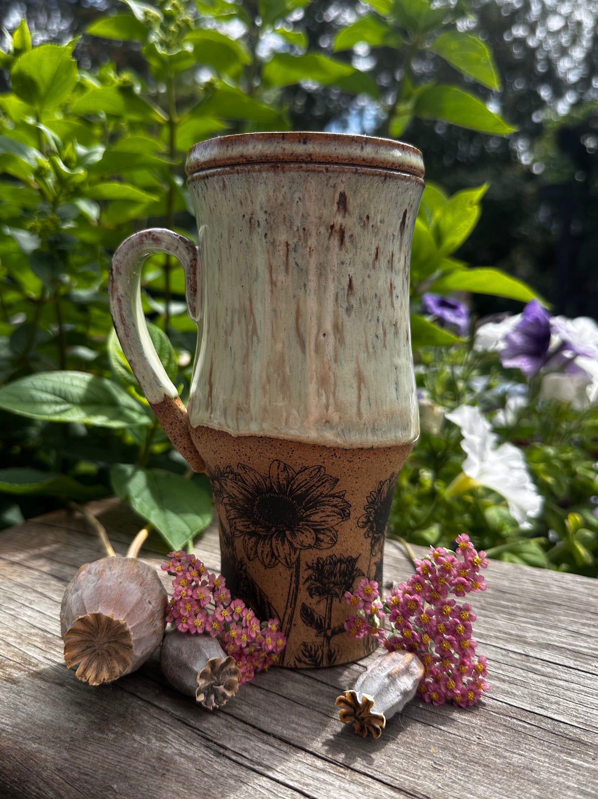 A rustic ceramic travel mug with a distressed cream and brown glaze, featuring a sunflower illustration on the lower half, displayed on a weathered wooden surface with dried flowers and green foliage in the background.