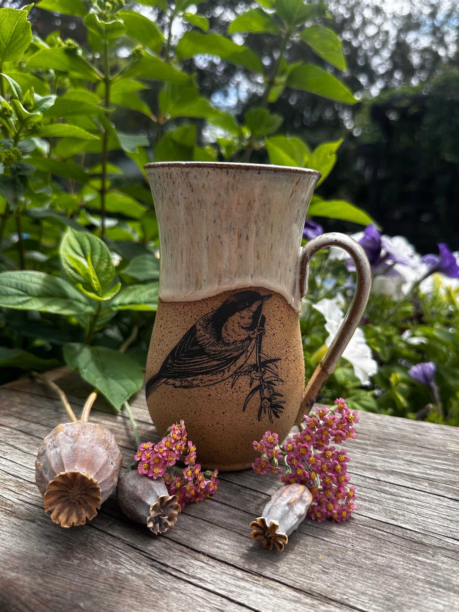 A handmade ceramic mug with a bird illustration, displayed on a weathered wooden surface with dried flowers and green foliage in the background.