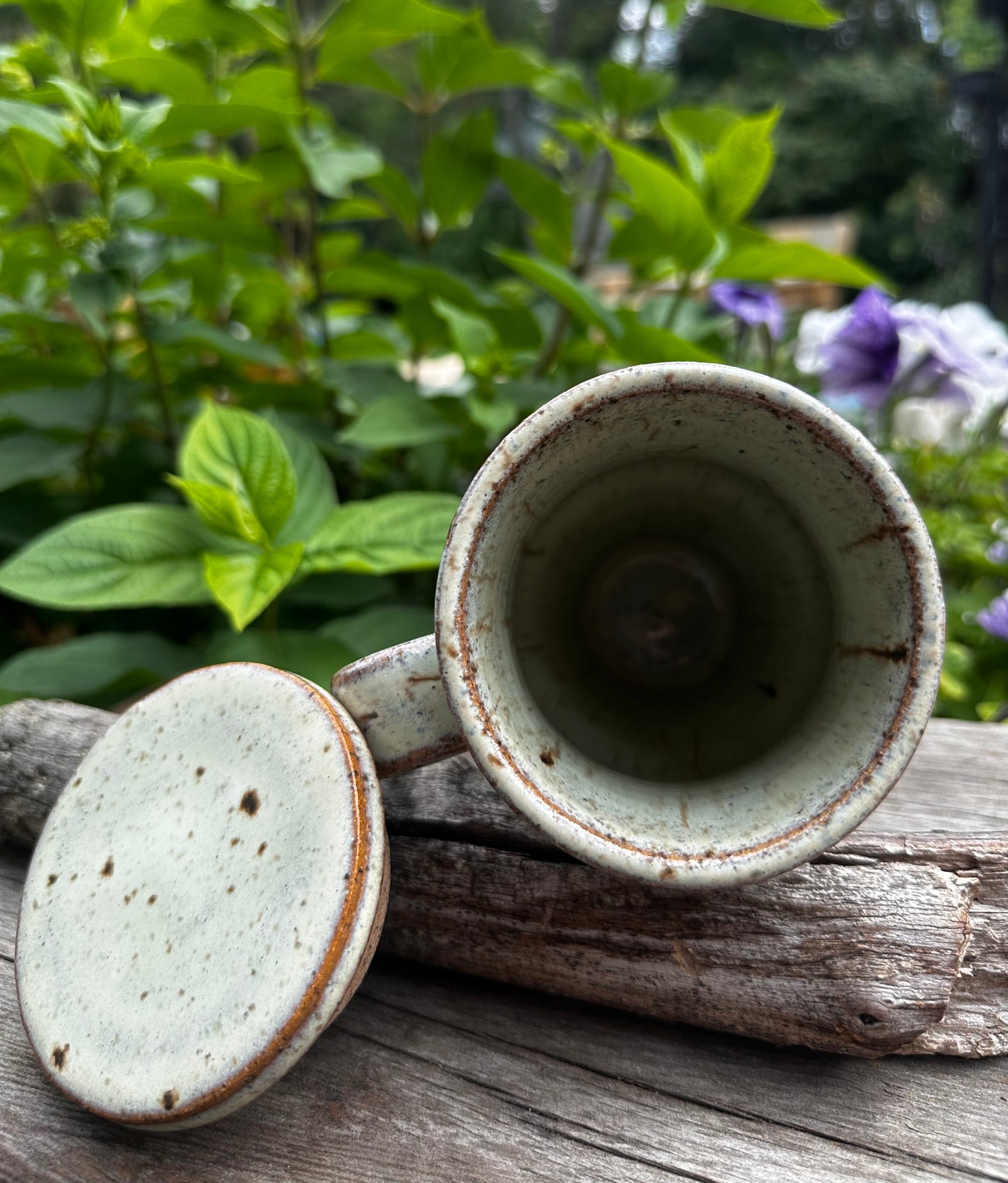 A handmade ceramic travel mug with a matching lid, displayed outdoors on a weathered log with green garden foliage in the background. The mug has a speckled green glaze with brown speckles and a thin brown rim, and the interior is a muted sage green.