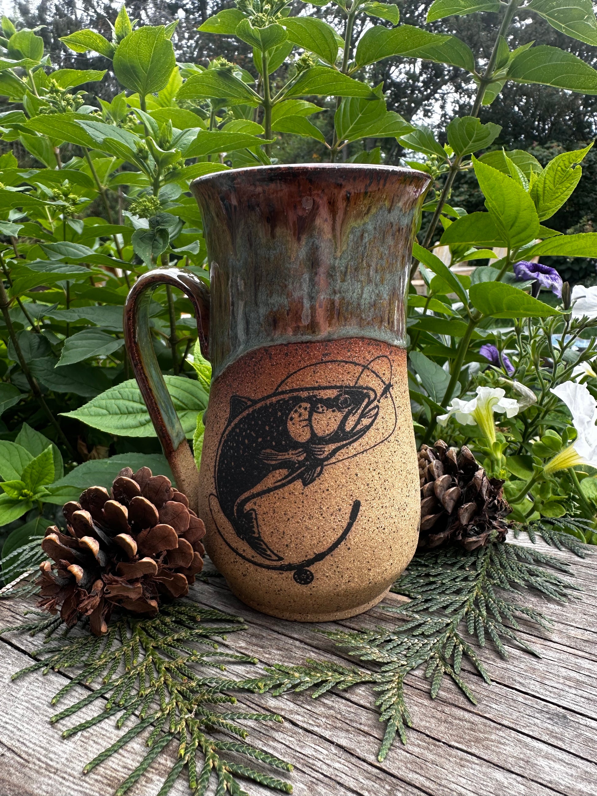 A rustic ceramic mug with a handle, decorated with a black fish design on a speckled tan body and a brown-green glazed rim, styled outdoors on a wooden surface with pinecones and greenery.