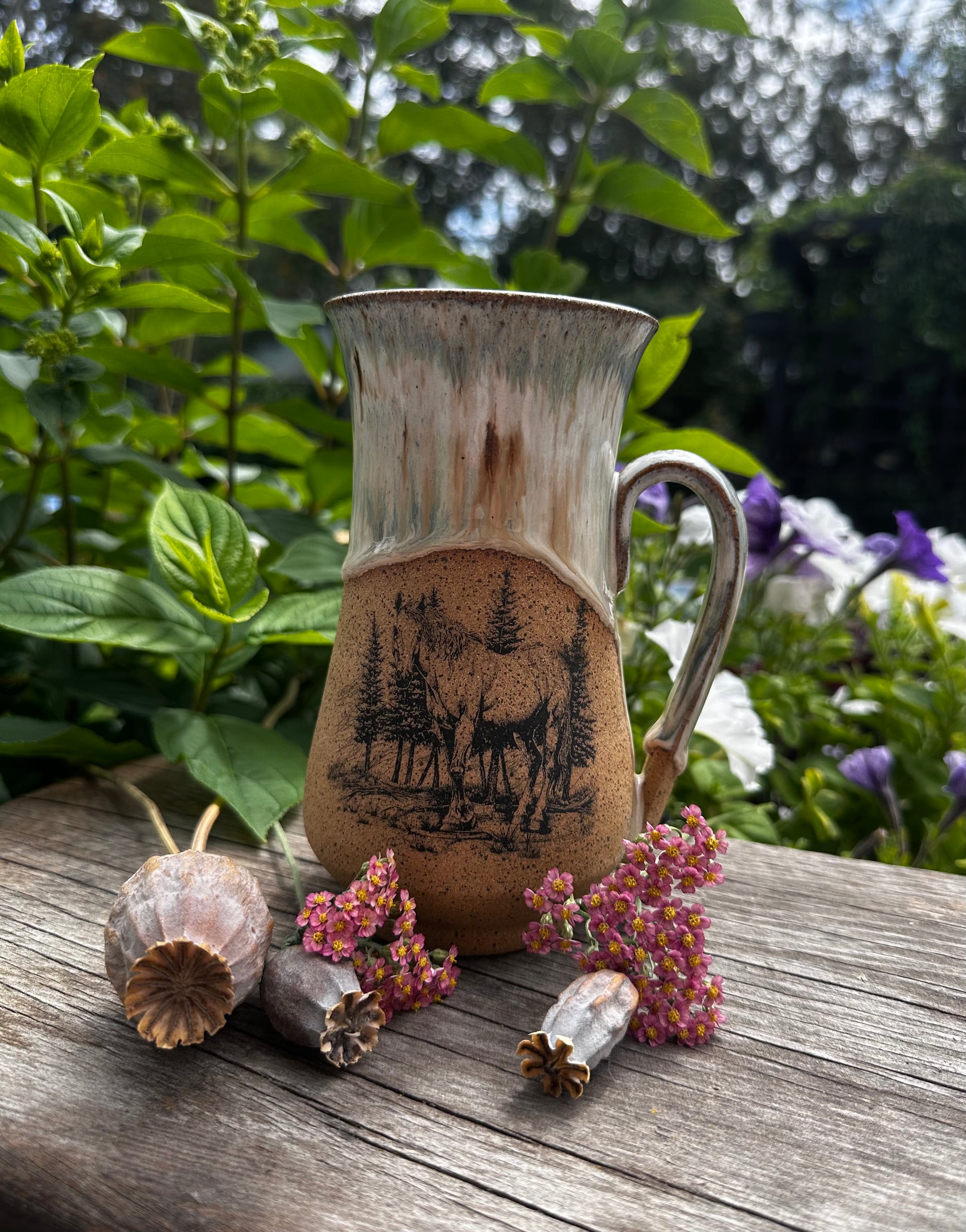 A rustic handmade ceramic mug with a handle, featuring a printed horse design on the front. The mug is placed on a weathered wooden surface outdoors, surrounded by dried poppy pods and small pink flowers, with green garden foliage in the background.