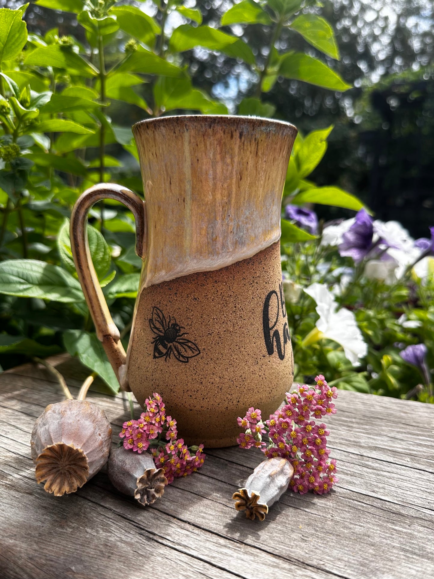 A rustic handmade ceramic mug with a handle, featuring a brown and beige glaze and a black bee illustration, set on a weathered wooden table with dried flowers and green garden foliage in the background.
