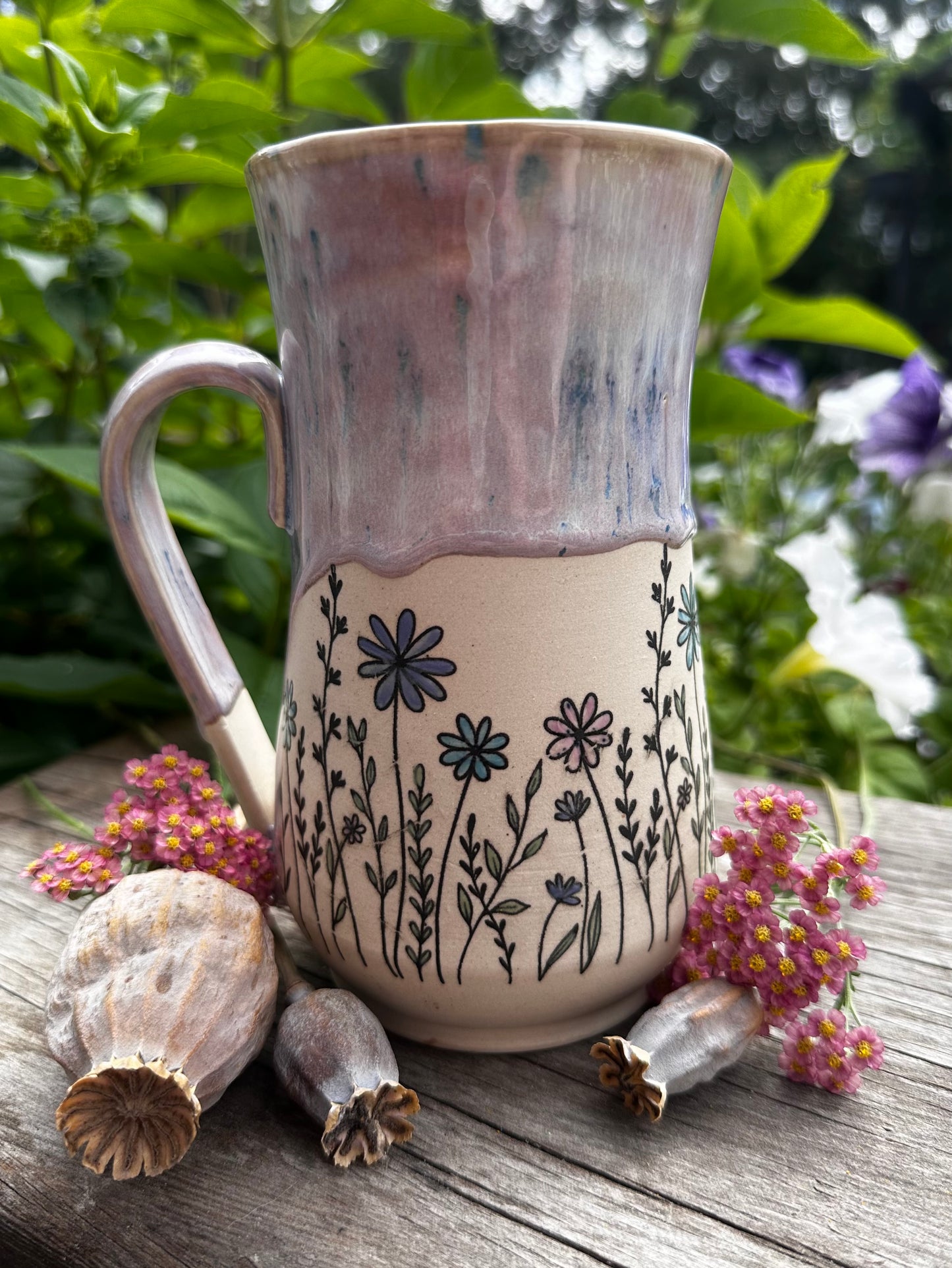 A handmade ceramic mug with a rustic dripping glaze and black floral line drawings, styled on a weathered wooden surface with dried poppy pods and pink flowers against a green garden background.