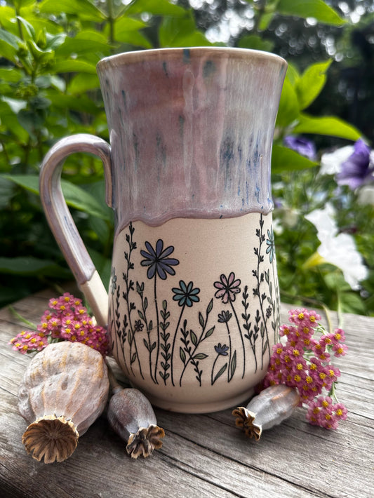 A handmade ceramic mug with a rustic dripping glaze and black floral line drawings, styled on a weathered wooden surface with dried poppy pods and pink flowers against a green garden background.