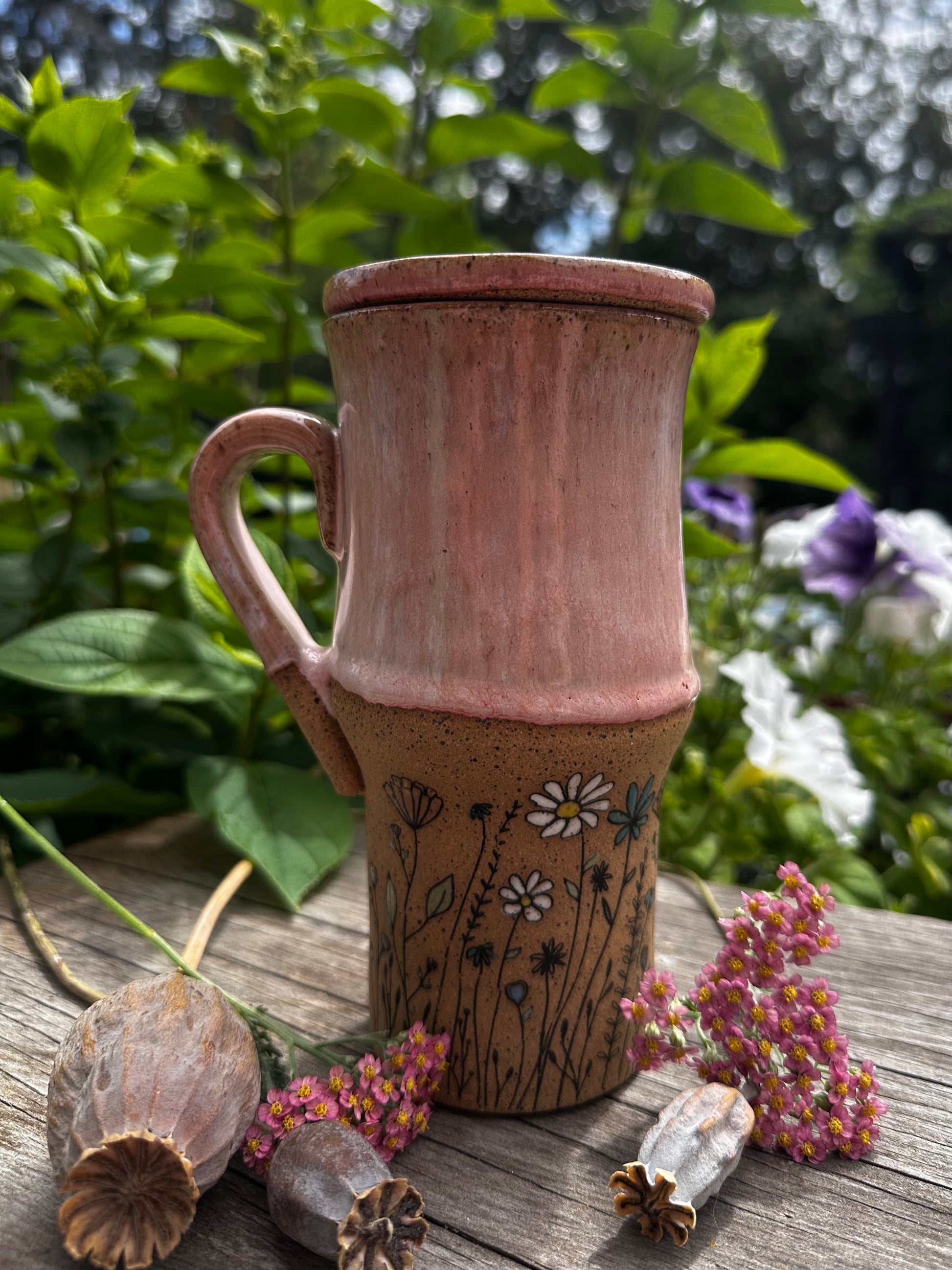 A handmade ceramic travel mug with a lid, featuring a pink upper section and a brown lower section decorated with black wildflower pattern. The mug is styled on a weathered wooden surface with dried poppy pods and small pink flowers, set against a soft-focus background of green garden foliage.
