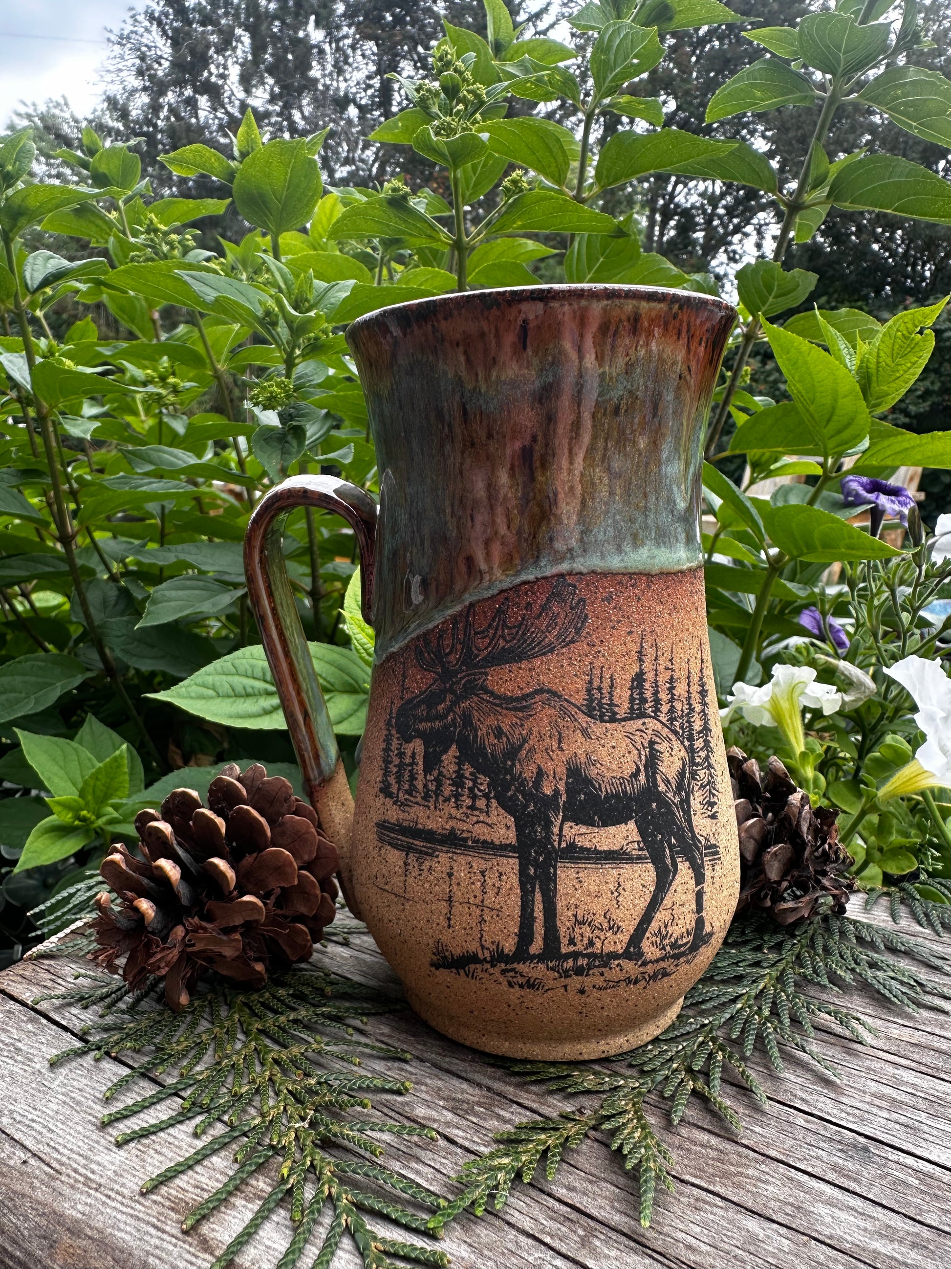 A rustic ceramic mug with a handle, featuring a brown and green glaze and a black moose illustration, styled on a wooden surface with pinecones and green foliage.