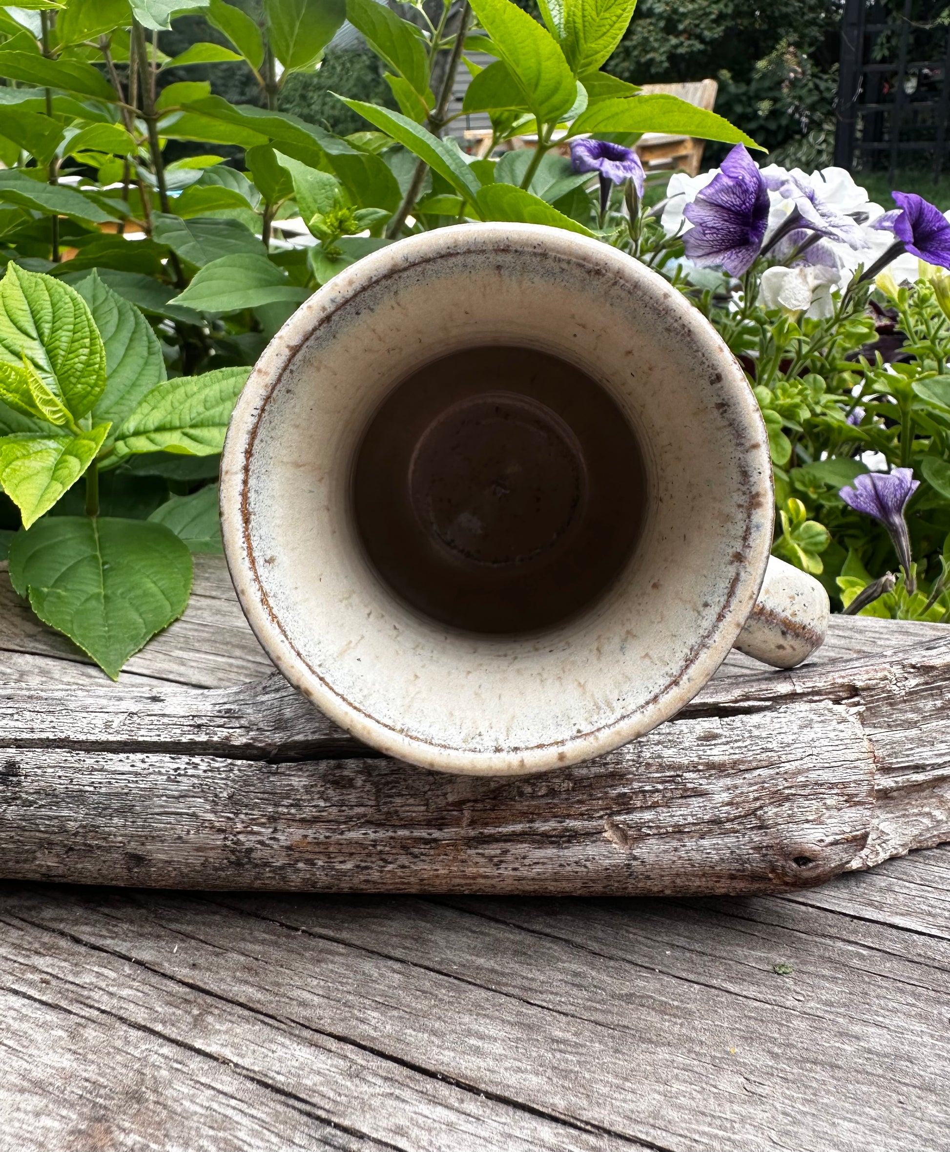 A handmade ceramic mug with a speckled beige glaze, resting on a weathered wooden log with green garden foliage and purple flowers in the background.