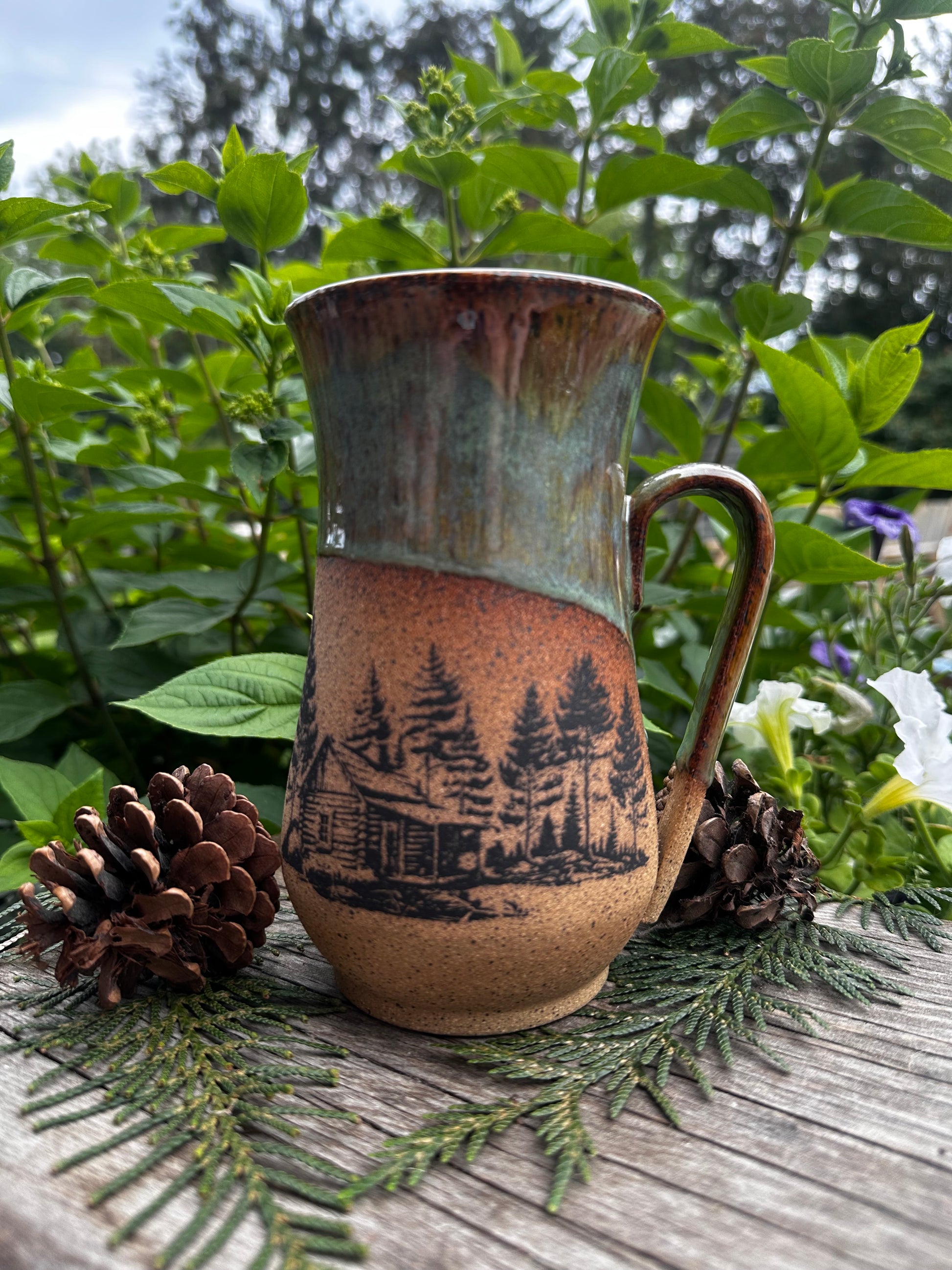A rustic ceramic mug with a handle, featuring a brown and green glaze and a black printed forest cabin design, styled on a wooden surface with pinecones and greenery.