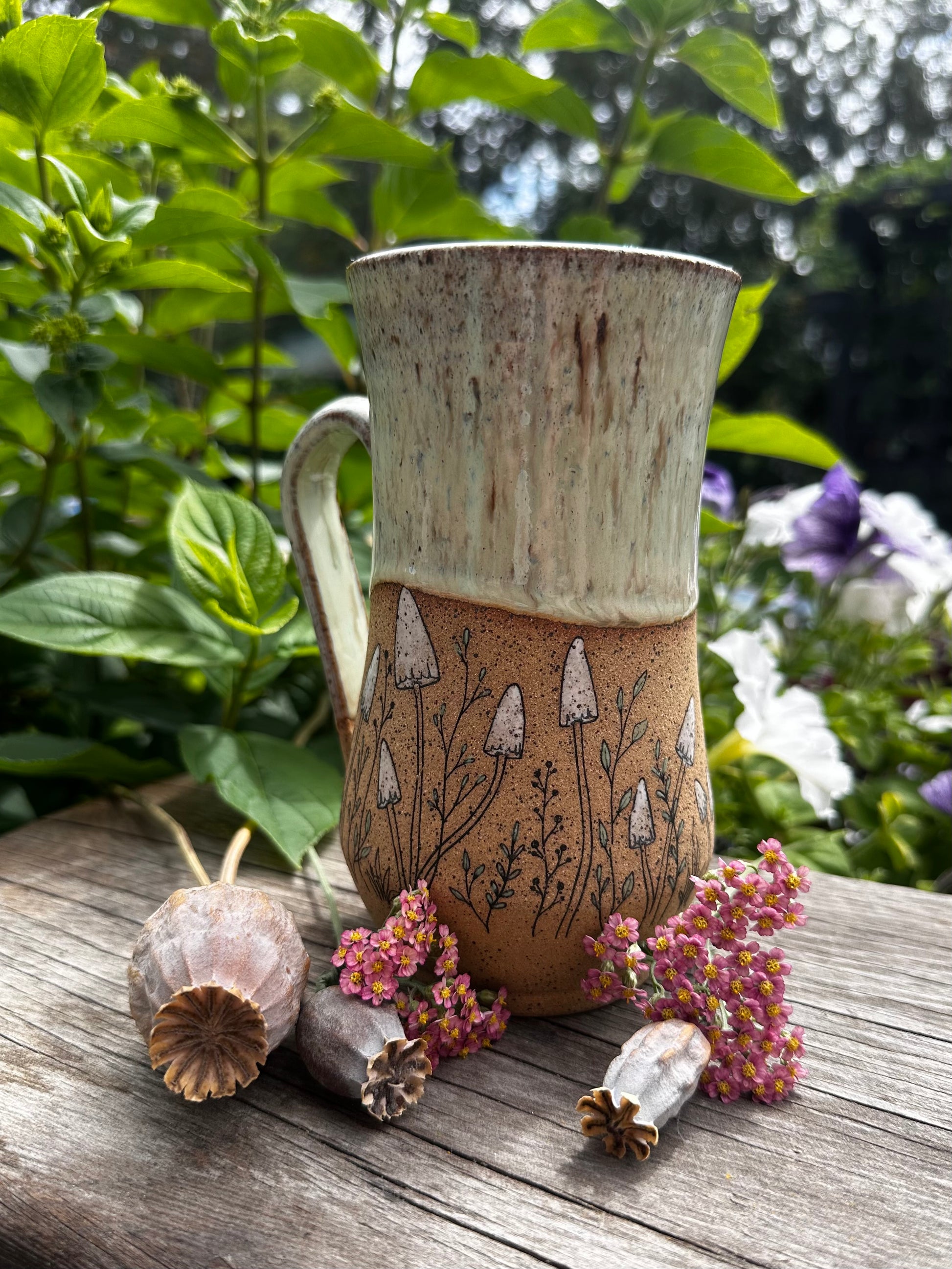 A handmade ceramic mug with a textured green and brown glaze, decorated with a hand-drawn style mushroom pattern. It is styled on a weathered wooden surface outdoors with green foliage and dried botanicals in the background