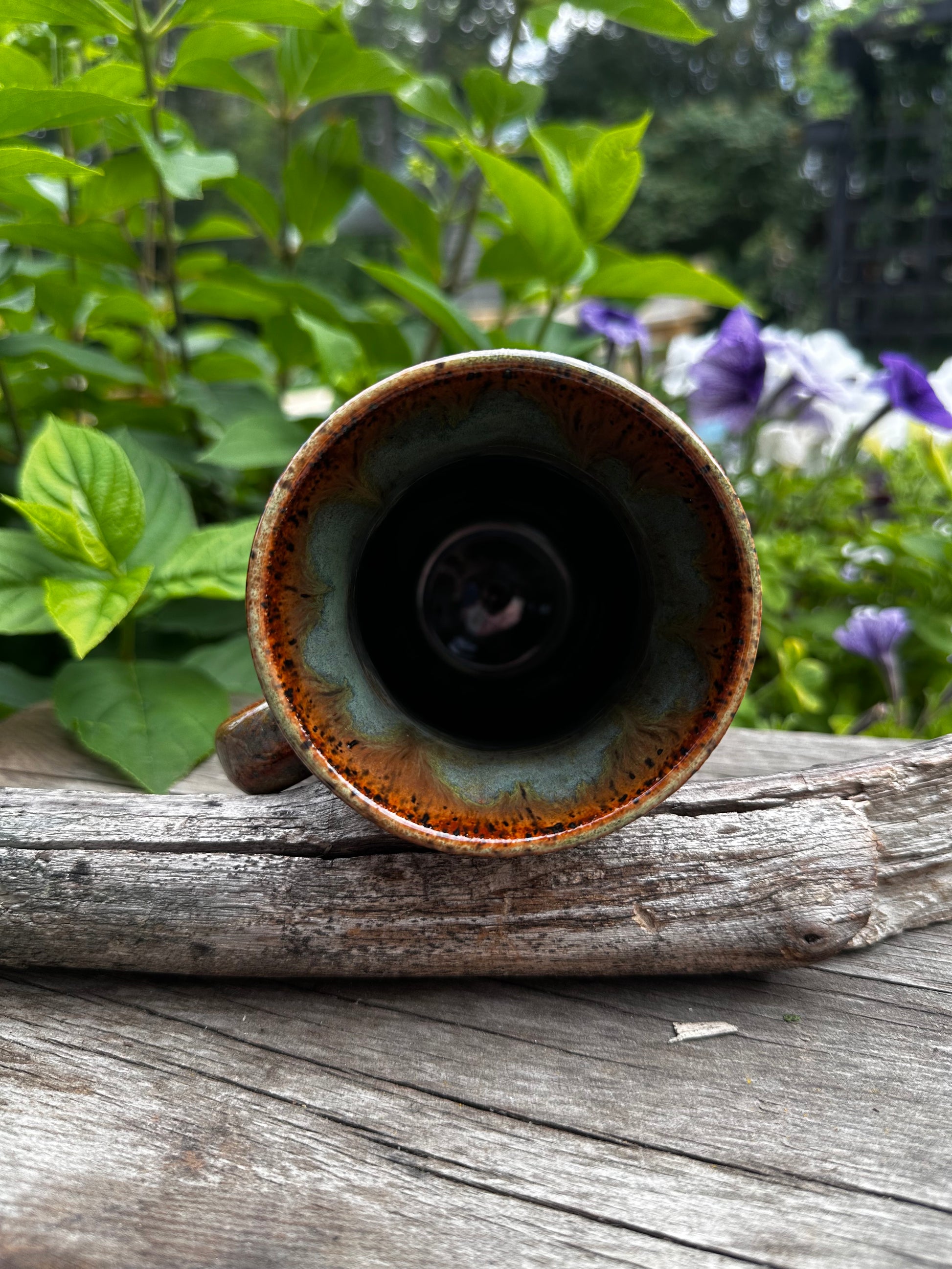 A handmade ceramic mug with a brown and green glaze, mounted on a weathered wooden post outdoors with green foliage and purple flowers in the background.