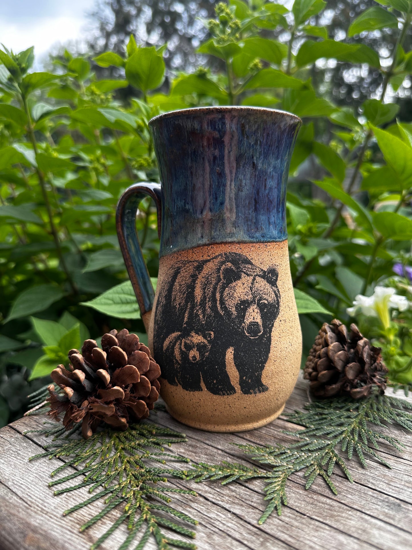 A handmade ceramic mug with a bear illustration, styled outdoors on a wooden surface with pinecones and greenery.