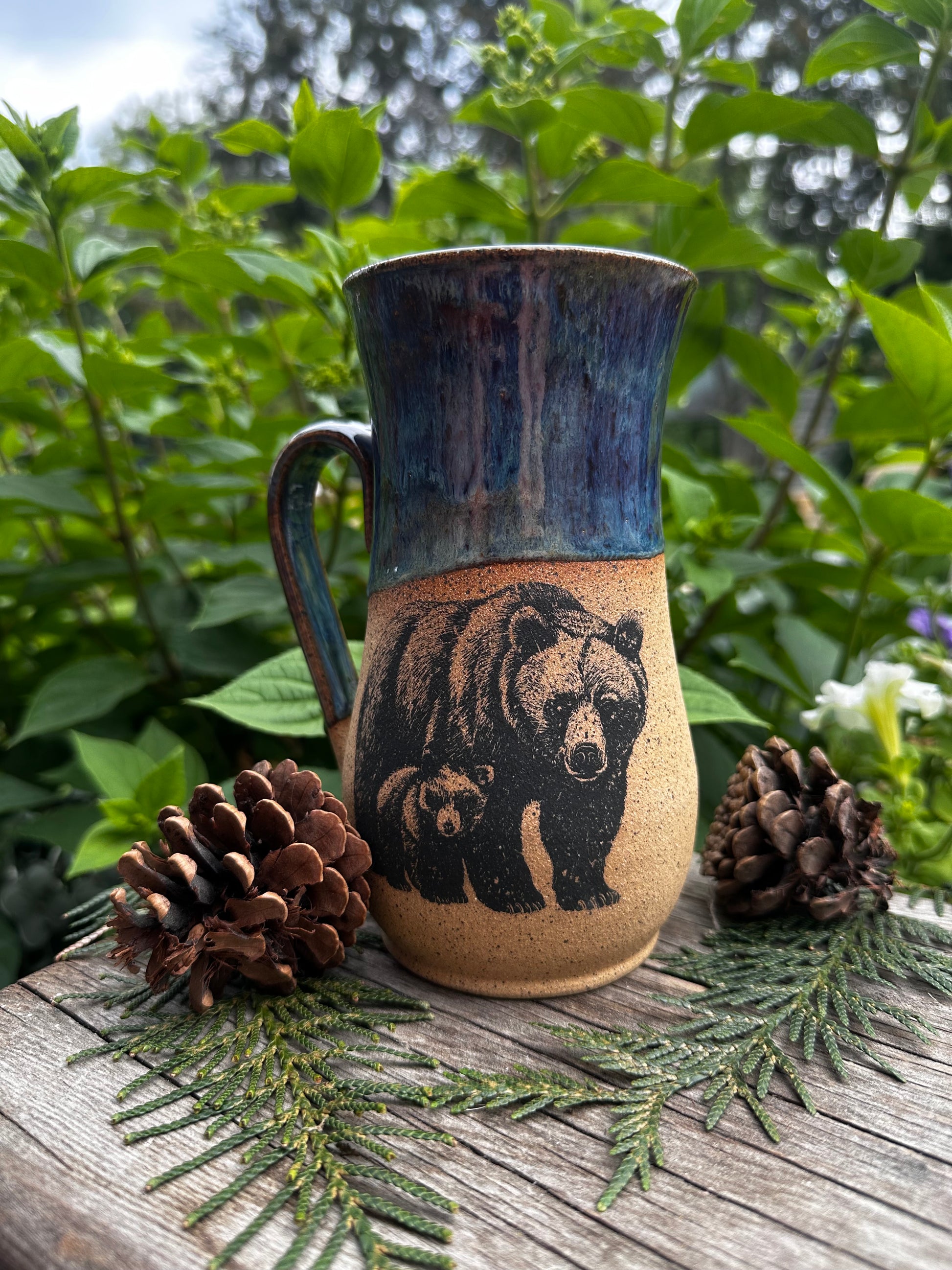 A handmade ceramic mug with a bear illustration, styled outdoors on a wooden surface with pinecones and greenery.