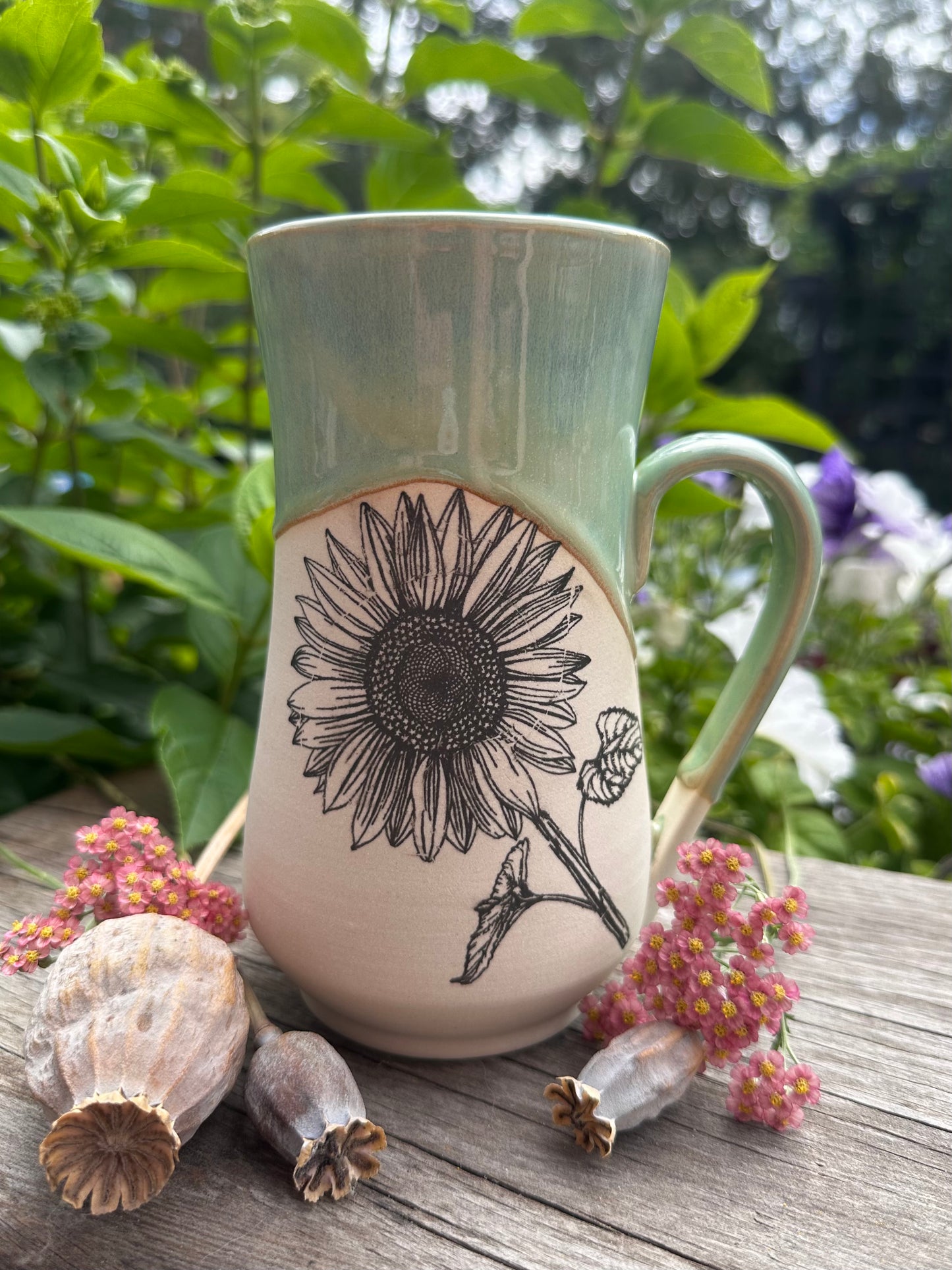 A ceramic mug with a green glazed upper half and a beige lower half featuring a black sunflower illustration, set on a wooden surface with dried flowers and green foliage in the background.