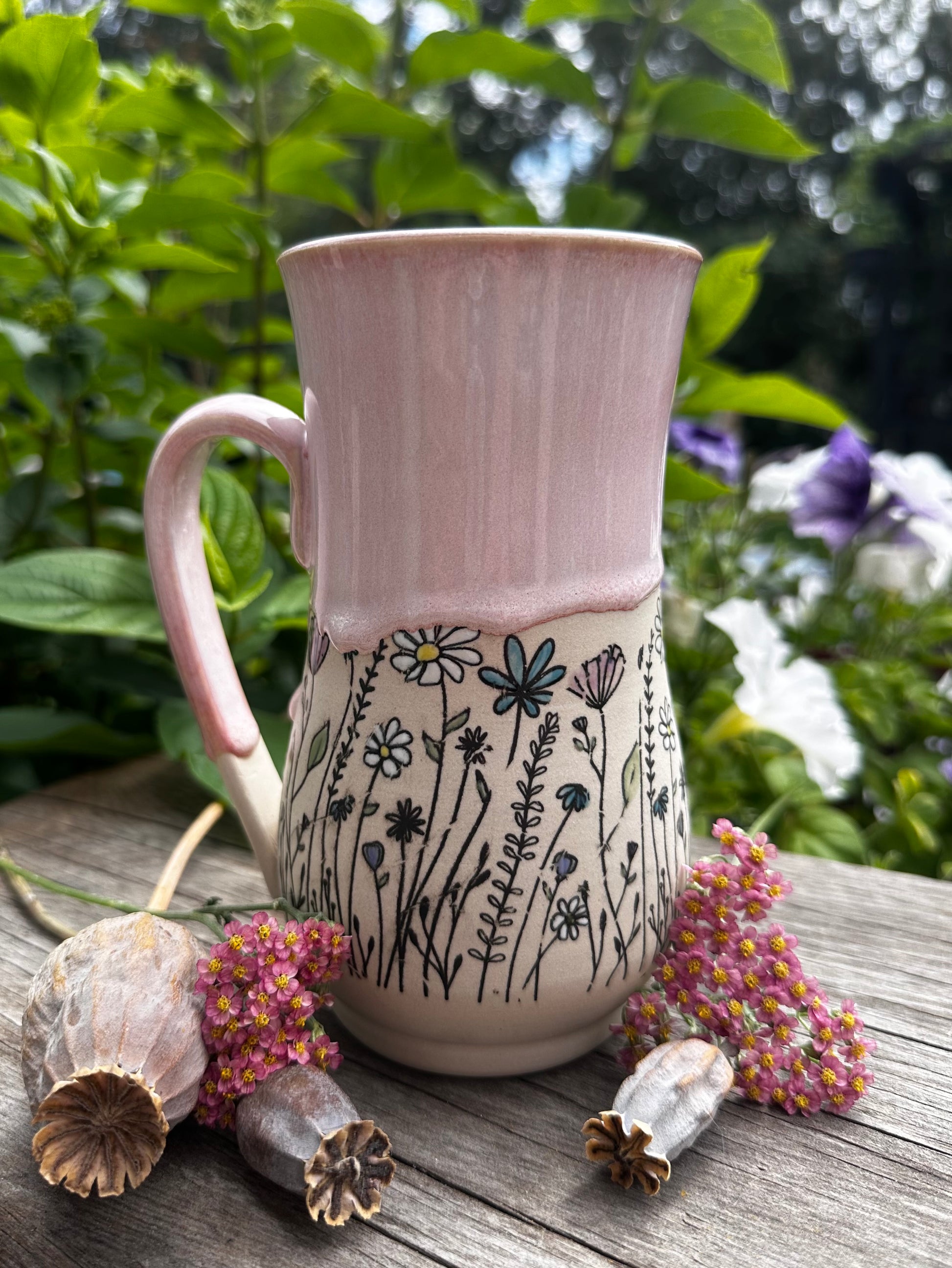 A handmade ceramic mug with a pink upper half and a cream lower half decorated with black wildflower illustrations, set on a wooden table outdoors with green foliage and dried flowers in the background.