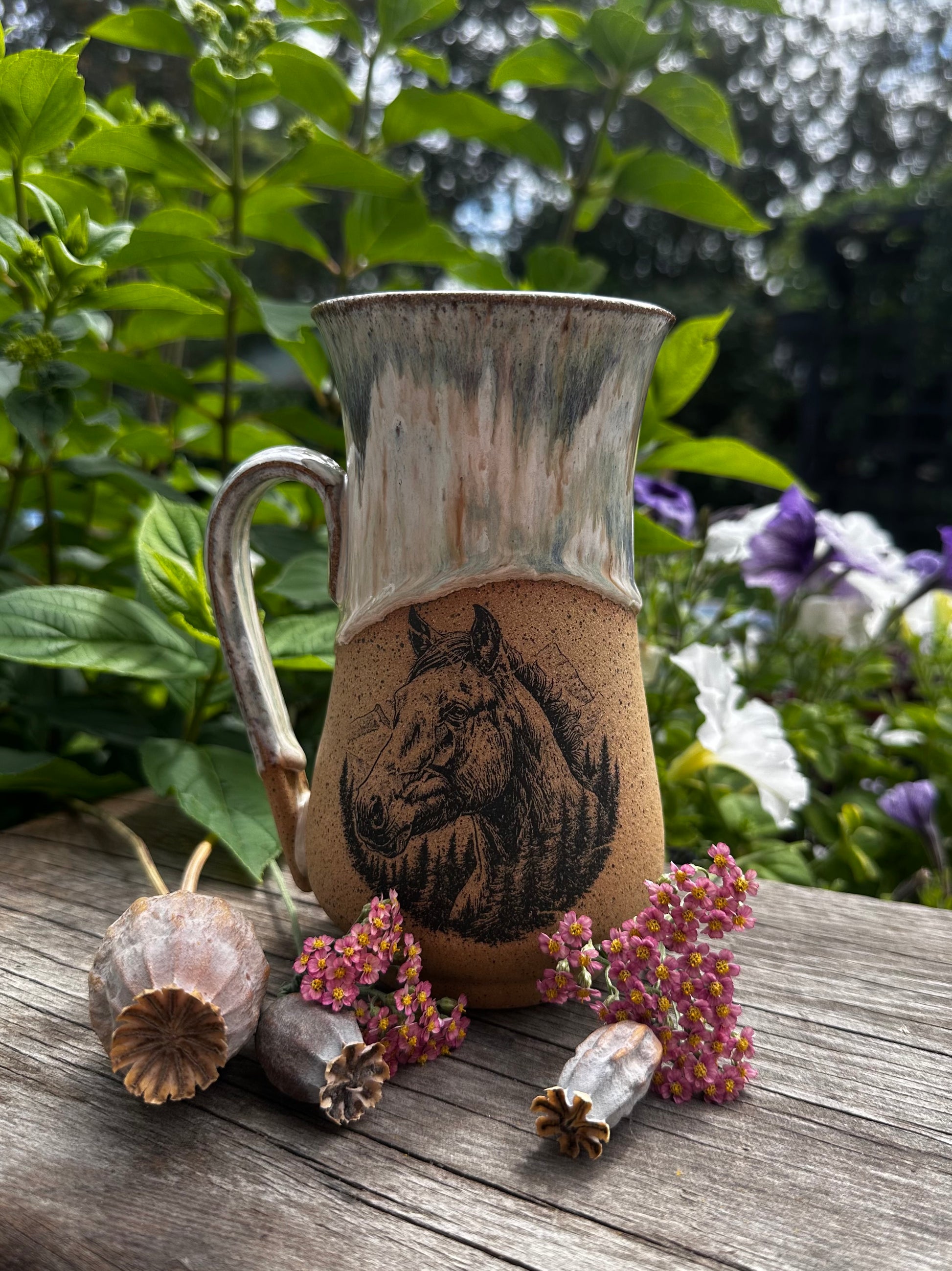 A rustic handmade ceramic mug with a handle, featuring a printed horse design on the front. The mug is placed on a weathered wooden surface outdoors, surrounded by dried poppy pods and small pink flowers, with green garden foliage in the background.