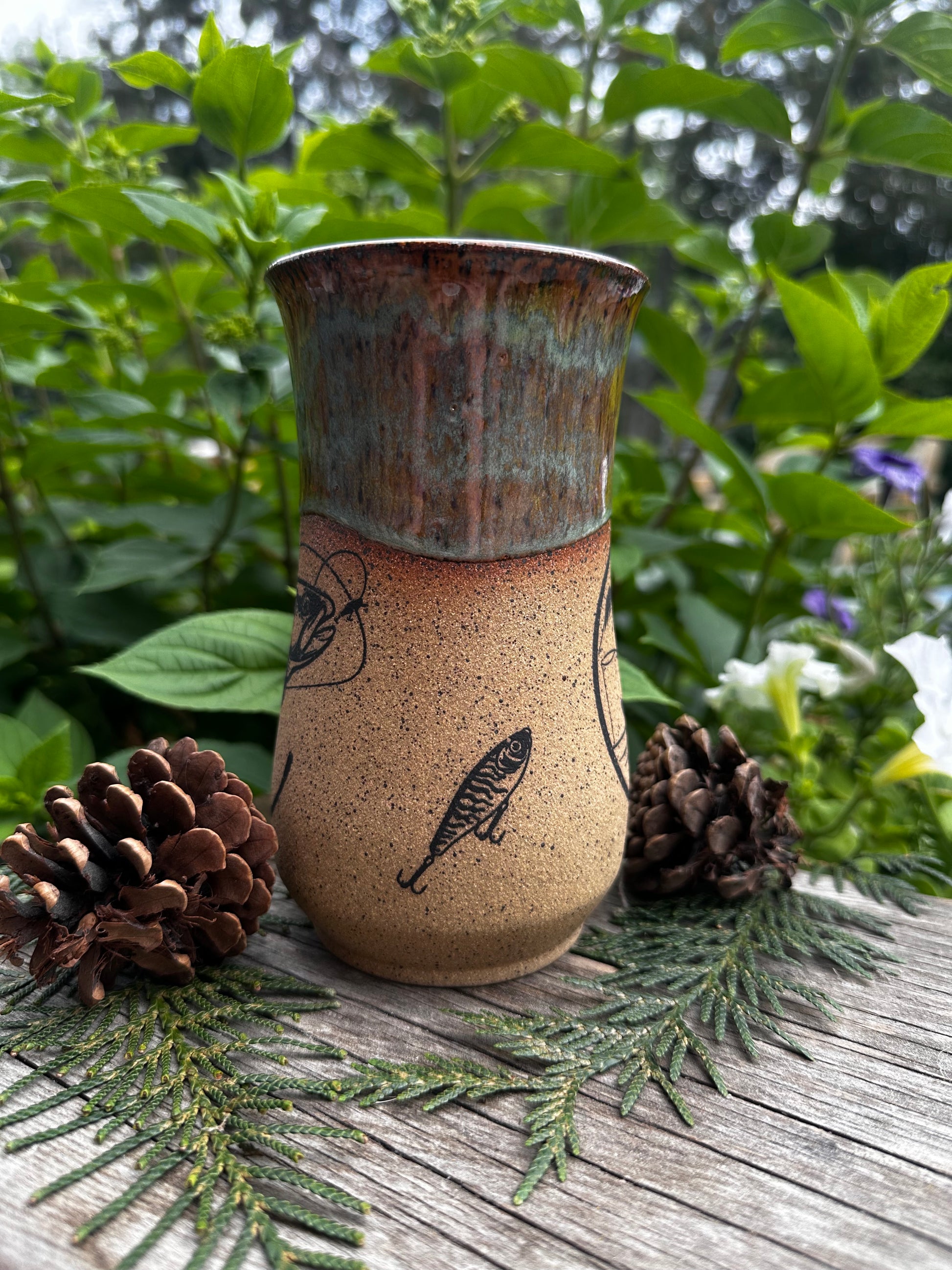 A handmade ceramic mug with a textured speckled beige body, a dark brown fish illustration, and a rustic greenish-brown glazed rim, styled on a wooden surface with pinecones and green foliage.