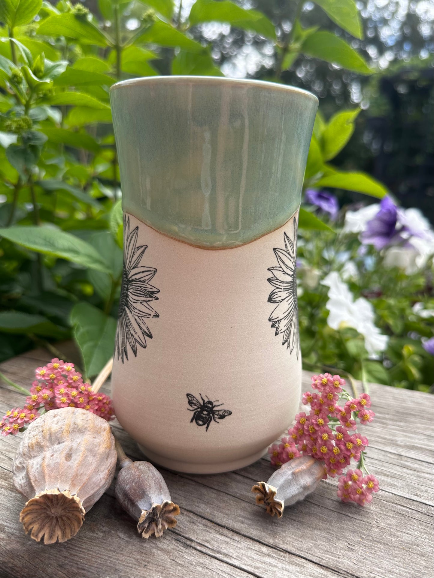 A handmade ceramic mug with a sage green glazed top and a cream-colored body decorated with black floral and bee illustrations, displayed on a rustic wooden surface with dried flowers and green garden foliage in the background.