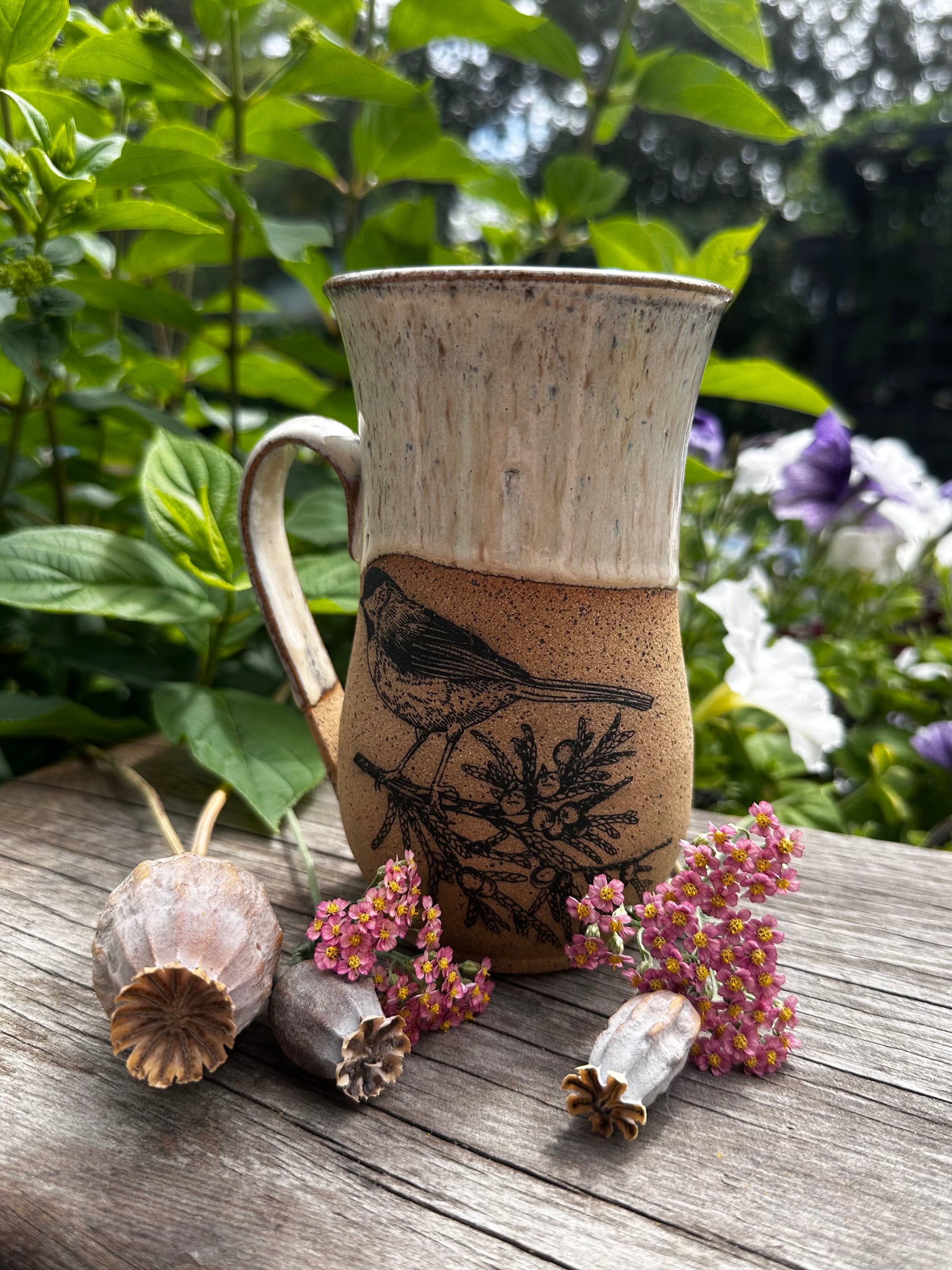 A handmade ceramic mug with a bird illustration, displayed on a weathered wooden surface with dried flowers and green foliage in the background.