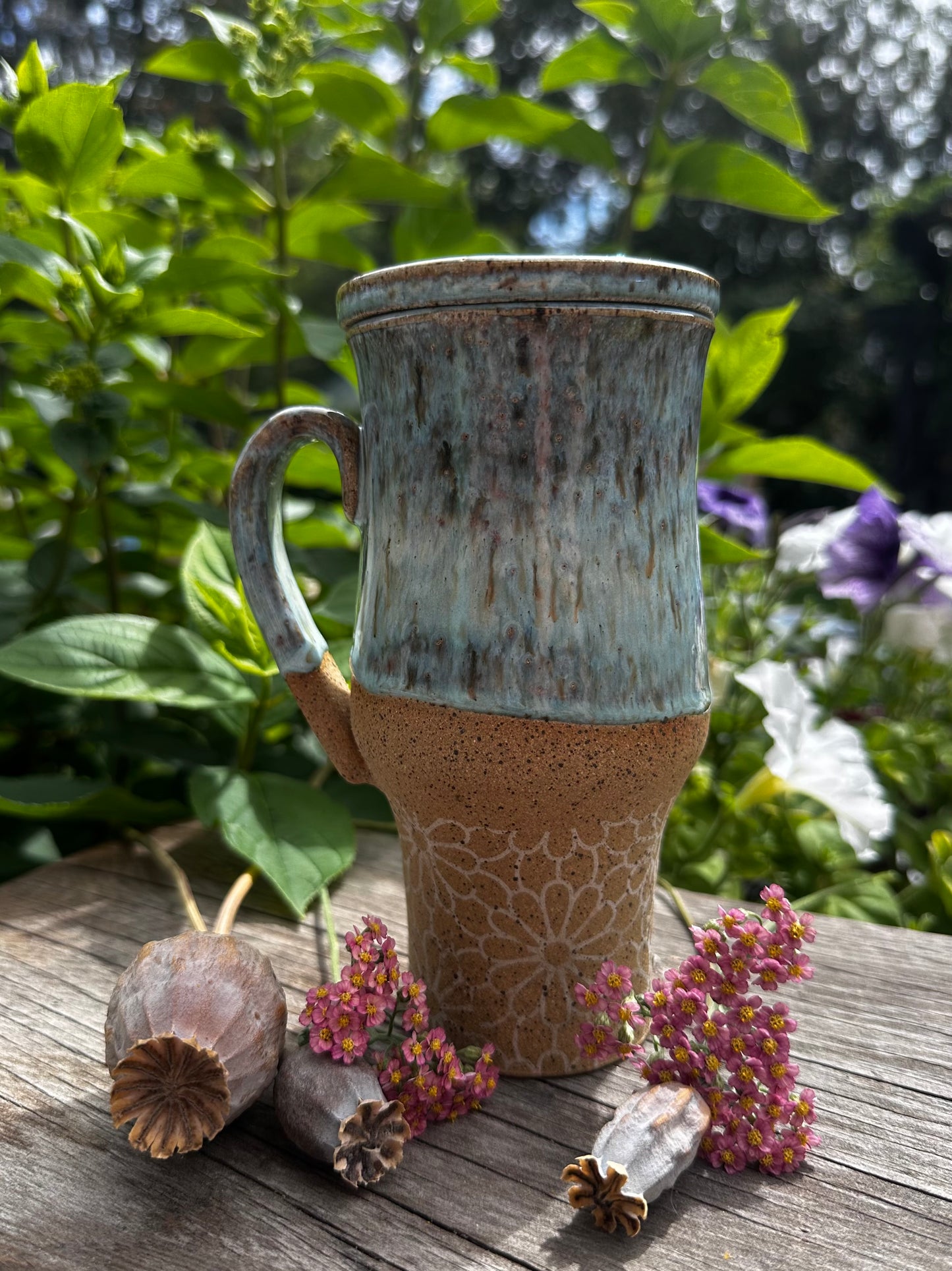 A handmade ceramic travel mug with a textured, speckled glaze sits on a rustic wooden table outdoors, surrounded by green foliage, purple flowers, and dried botanicals. The mug features a light blue glazed upper half and a natural, speckled tan-brown lower half with a floral pattern.