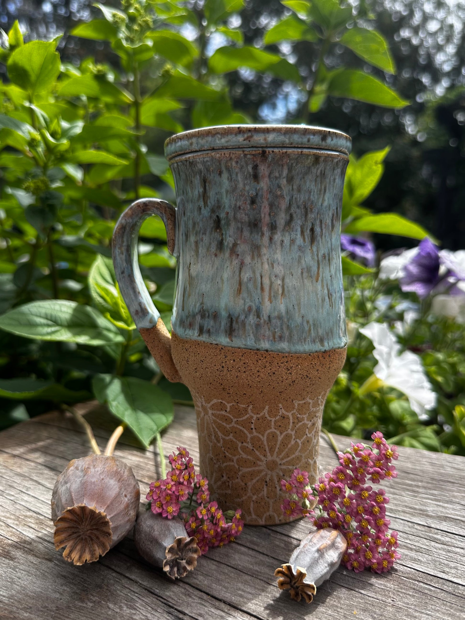 A handmade ceramic travel mug with a textured, speckled glaze sits on a rustic wooden table outdoors, surrounded by green foliage, purple flowers, and dried botanicals. The mug features a light blue glazed upper half and a natural, speckled tan-brown lower half with a floral pattern.