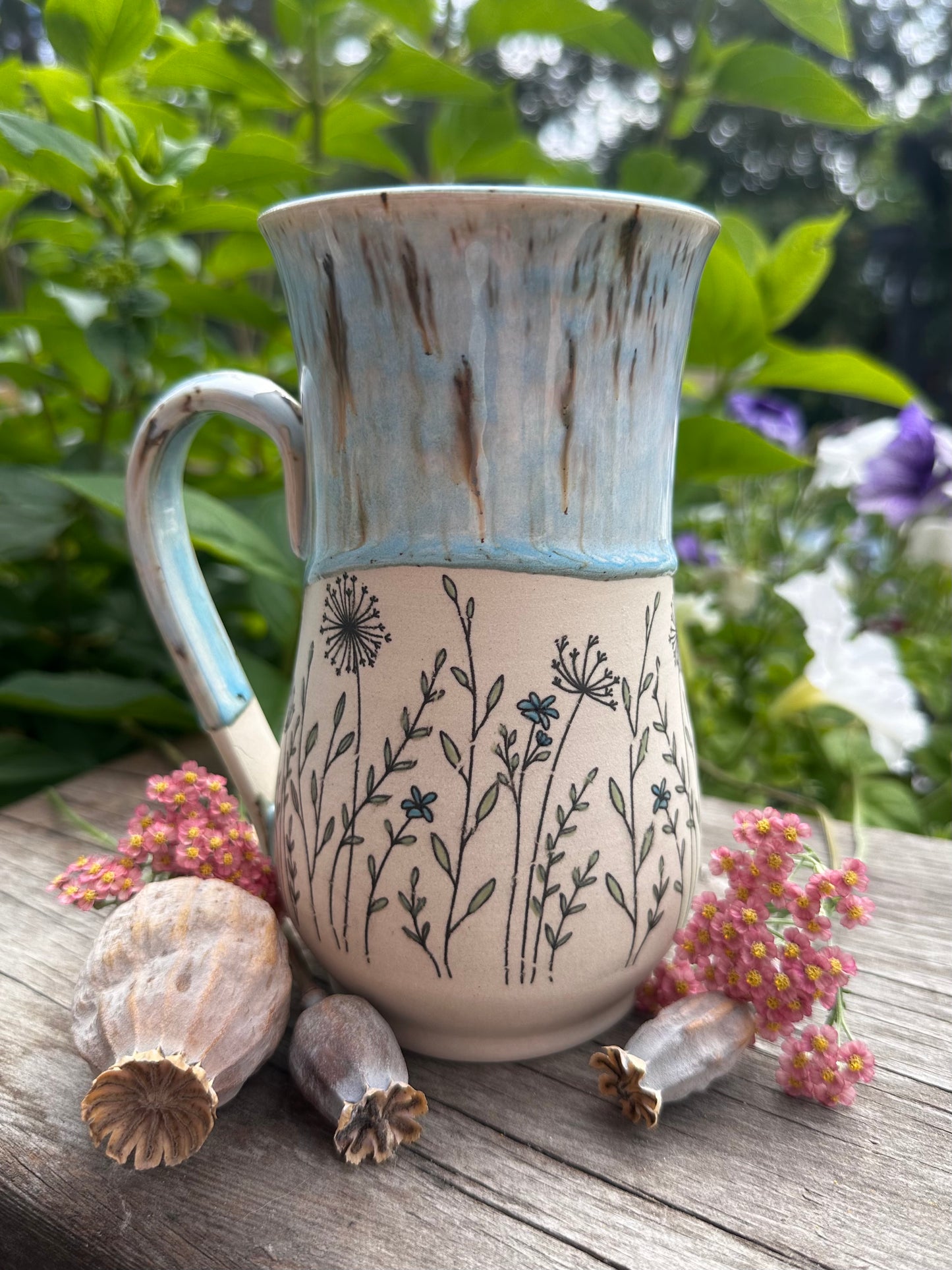 A handmade ceramic mug with a light blue glazed upper half and a cream lower half decorated with black wildflower illustrations, displayed on a rustic wooden surface with dried flowers and green foliage in the background.
