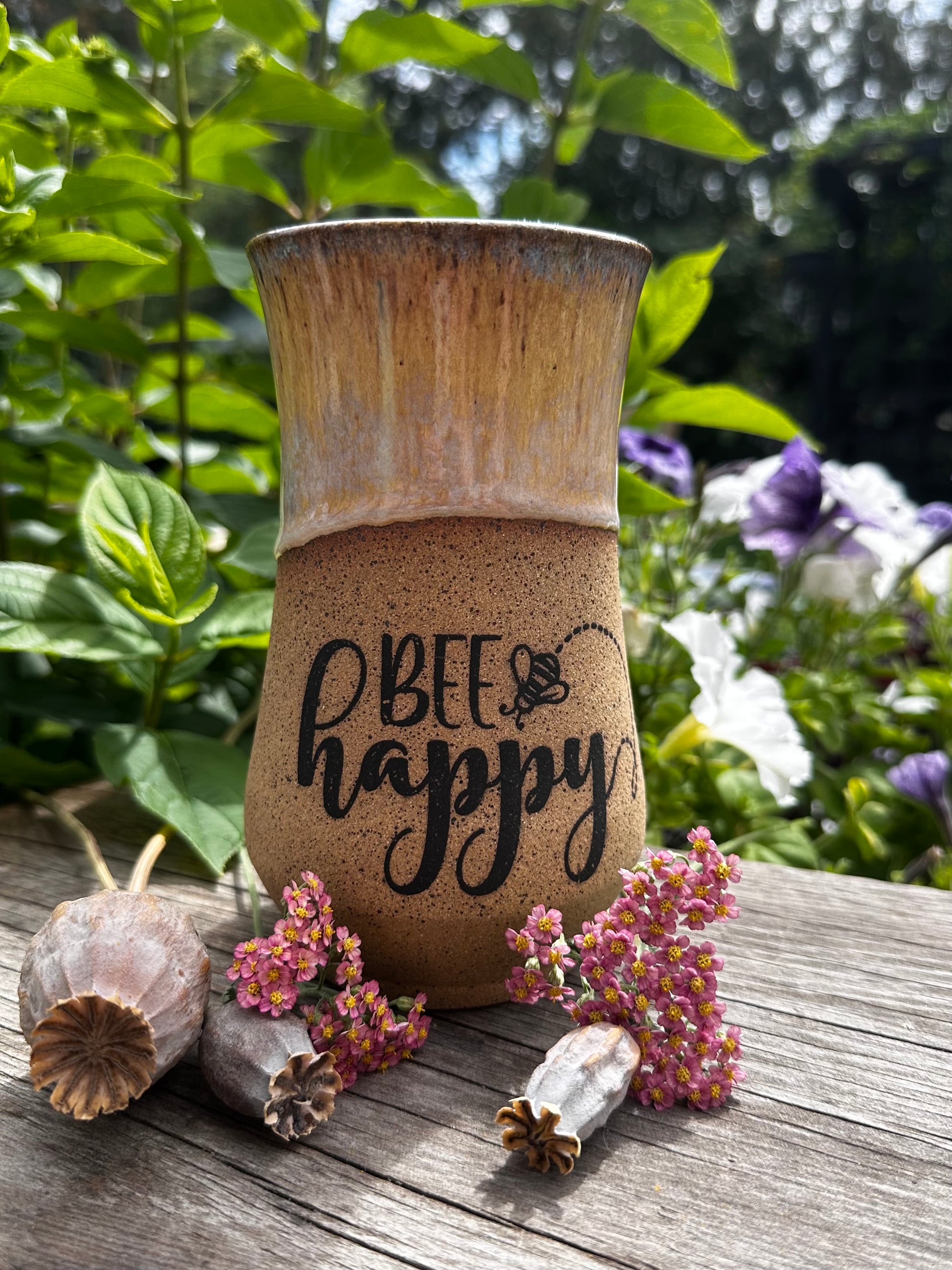 A rustic ceramic mug with a two-tone brown and beige glaze, featuring the text 'BEE happy' in black script. The mug is styled on a weathered wooden surface with dried botanicals, set against a soft-focus background of green garden foliage and purple flowers.