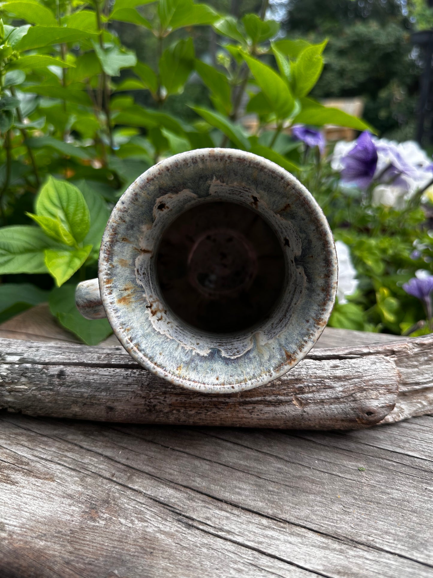 A rustic ceramic cup with a distressed, weathered glaze, shown from a top-down view resting against a weathered wooden post with green foliage and purple flowers in the background.
