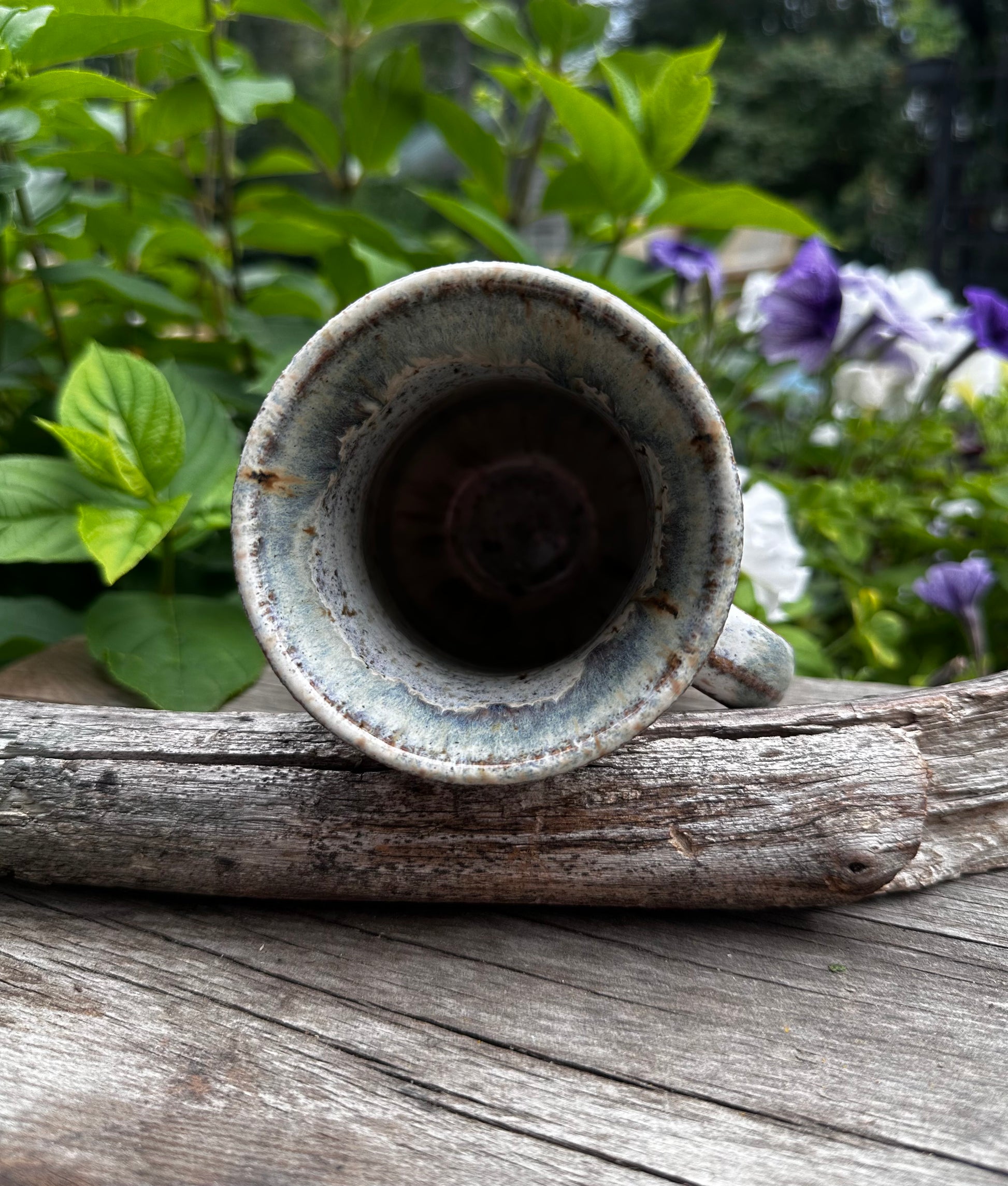 A rustic ceramic mug with a distressed blue-white glaze, resting on a weathered log with green garden foliage and purple flowers in the background.