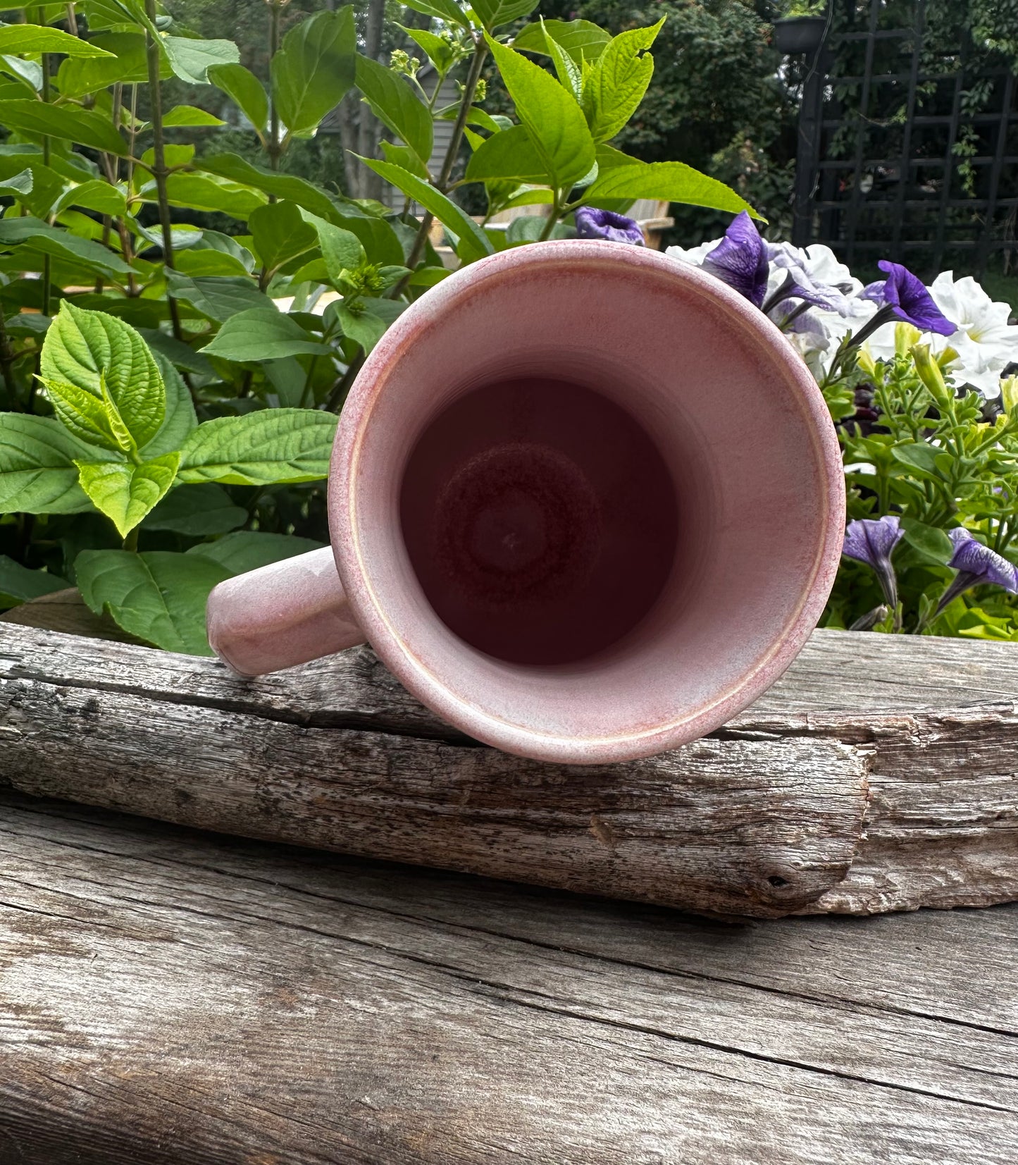 A pink ceramic mug with a handle, shown from a top-down perspective resting on a weathered wooden log with green garden foliage and purple flowers in the background.