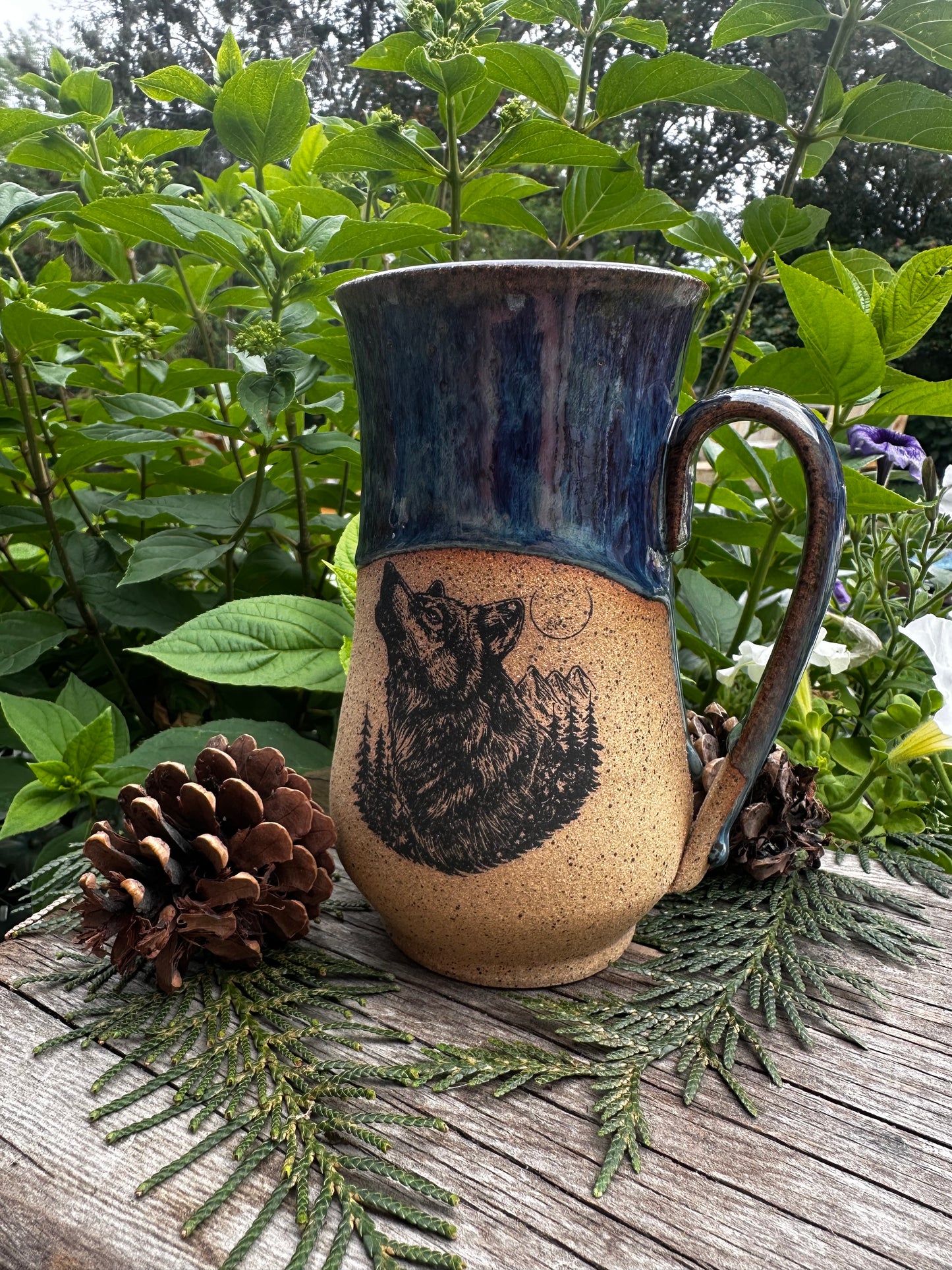 A handmade ceramic mug with a rustic handle, featuring a dark blue glazed upper section and a natural beige lower section with a black printed wolf design. The mug is styled on a weathered wooden surface surrounded by green pine branches, pinecones, and lush green foliage.