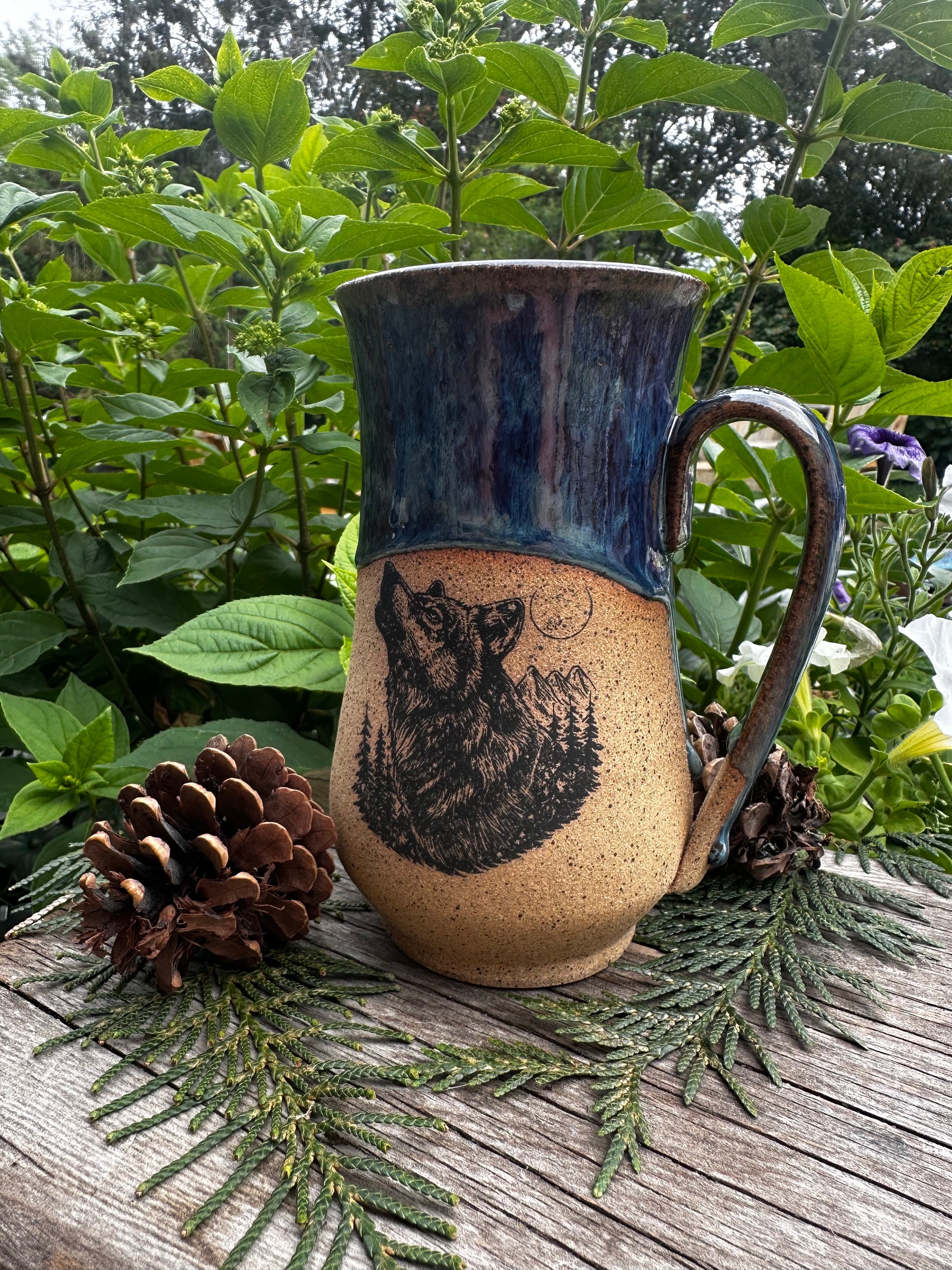 A handmade ceramic mug with a rustic handle, featuring a dark blue glazed upper section and a natural beige lower section with a black printed wolf design. The mug is styled on a weathered wooden surface surrounded by green pine branches, pinecones, and lush green foliage.