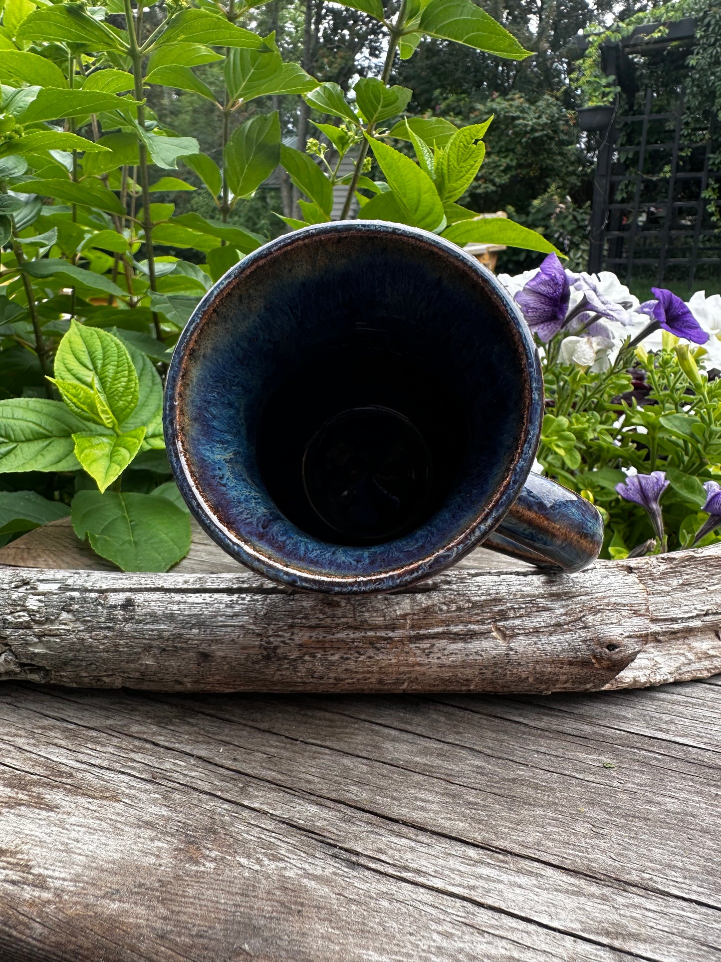 A handmade ceramic mug with a dark blue glaze, shown from a side angle resting on a weathered log outdoors with green foliage and purple flowers in the background.