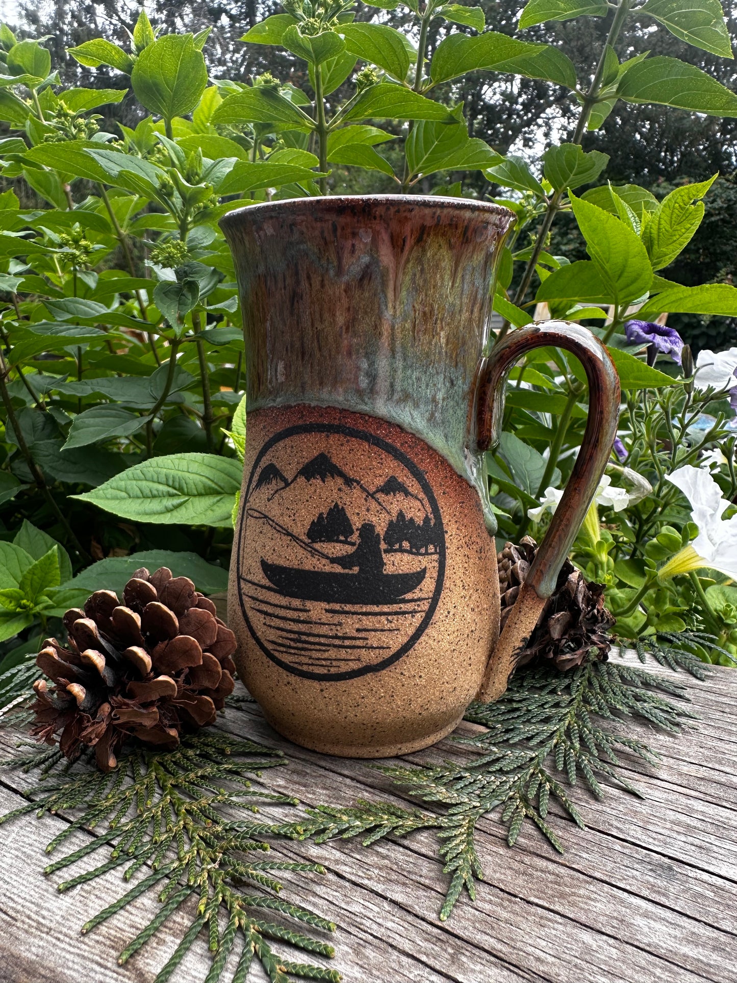 A handmade ceramic mug with a rustic brown and green glaze, featuring a black printed mountain and boat illustration. The mug is styled on a weathered wooden surface with pinecones and green foliage in the background.