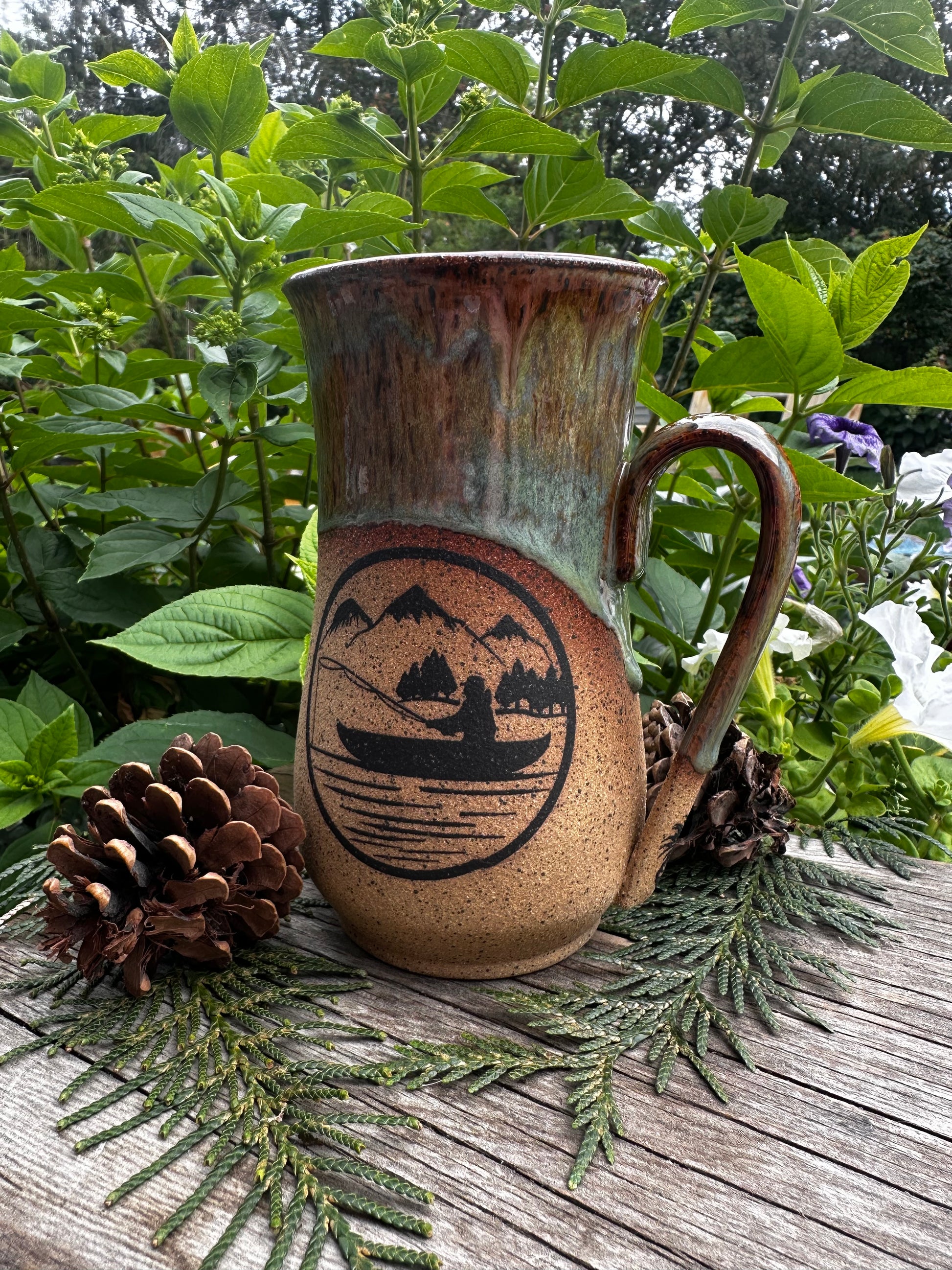 A handmade ceramic mug with a rustic brown and green glaze, featuring a black printed mountain and boat illustration. The mug is styled on a weathered wooden surface with pinecones and green foliage in the background.