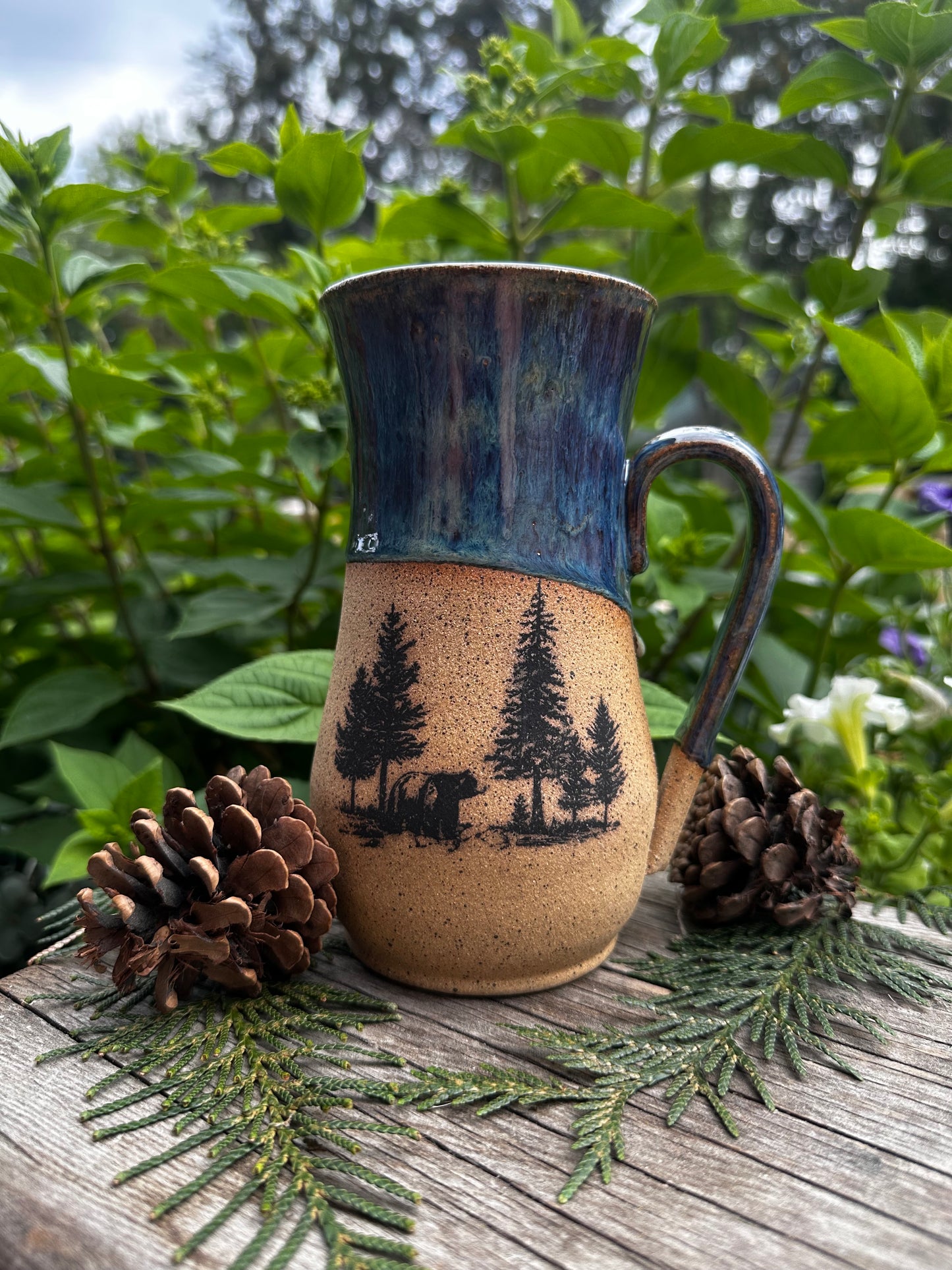 A handmade ceramic mug with a rustic handle, featuring a dark blue glazed upper half and a speckled beige lower half with a black tree silhouette design. The mug is styled outdoors on a wooden surface with pinecones and evergreen branches against a background of green foliage.
