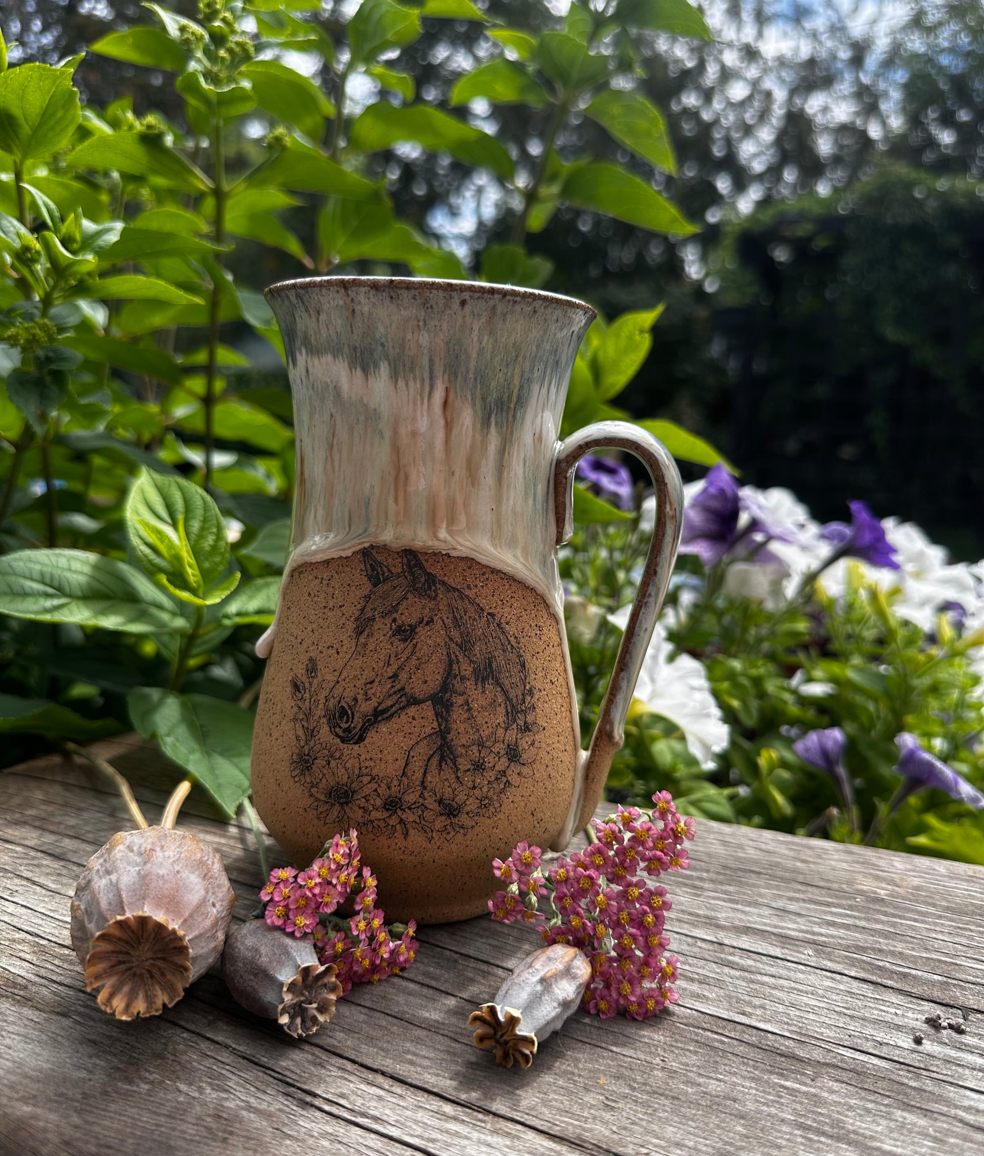 A handmade ceramic mug with a rustic finish, featuring a painted horse head design on the lower half. The mug is displayed on a weathered wooden table outdoors, surrounded by green foliage, purple flowers, and dried botanicals.
