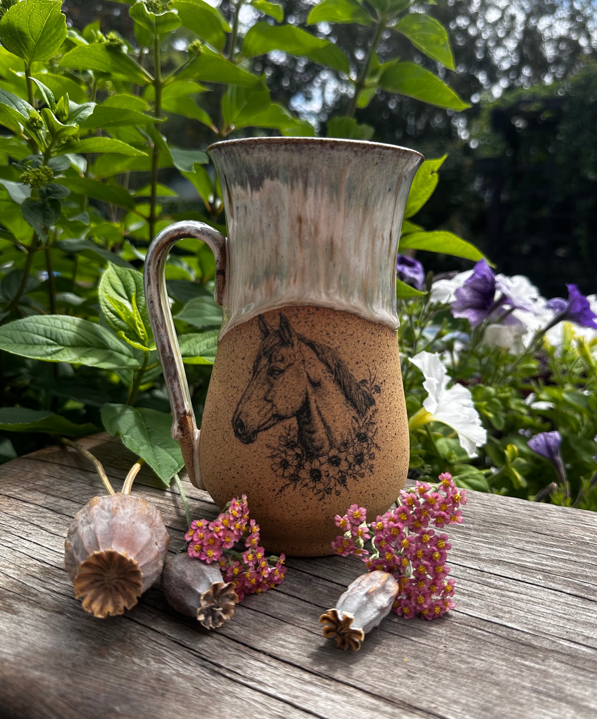 A rustic ceramic mug with a horse illustration, displayed on a weathered wooden surface outdoors with green foliage and small dried flowers in the background.