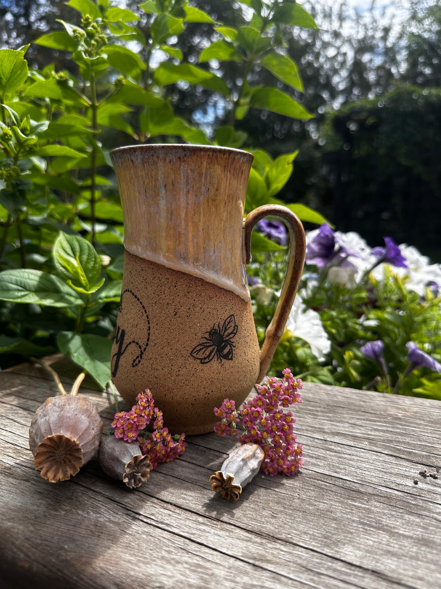 A rustic handmade ceramic mug with a handle, featuring a brown and beige glaze and a black bee illustration, set on a weathered wooden table with dried flowers and green garden foliage in the background.