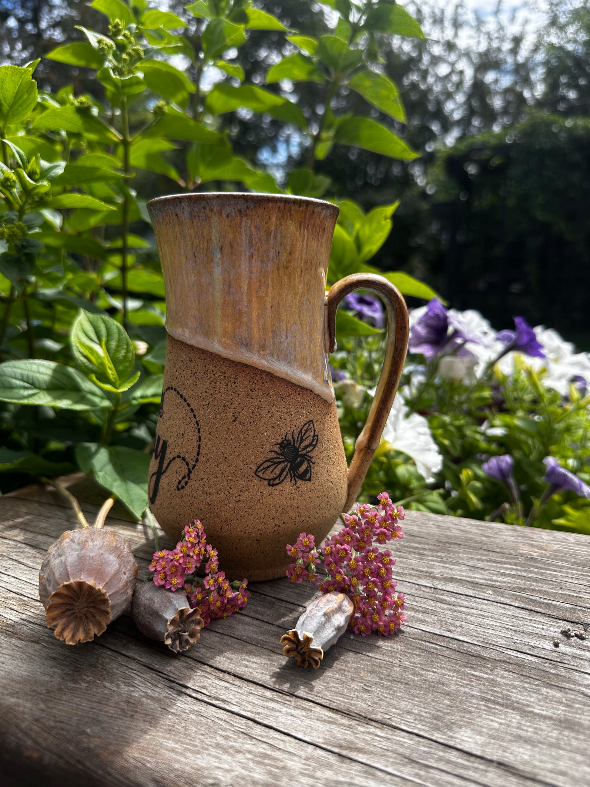 A rustic handmade ceramic mug with a handle, featuring a brown and beige glaze and a black bee illustration, set on a weathered wooden table with dried flowers and green garden foliage in the background.